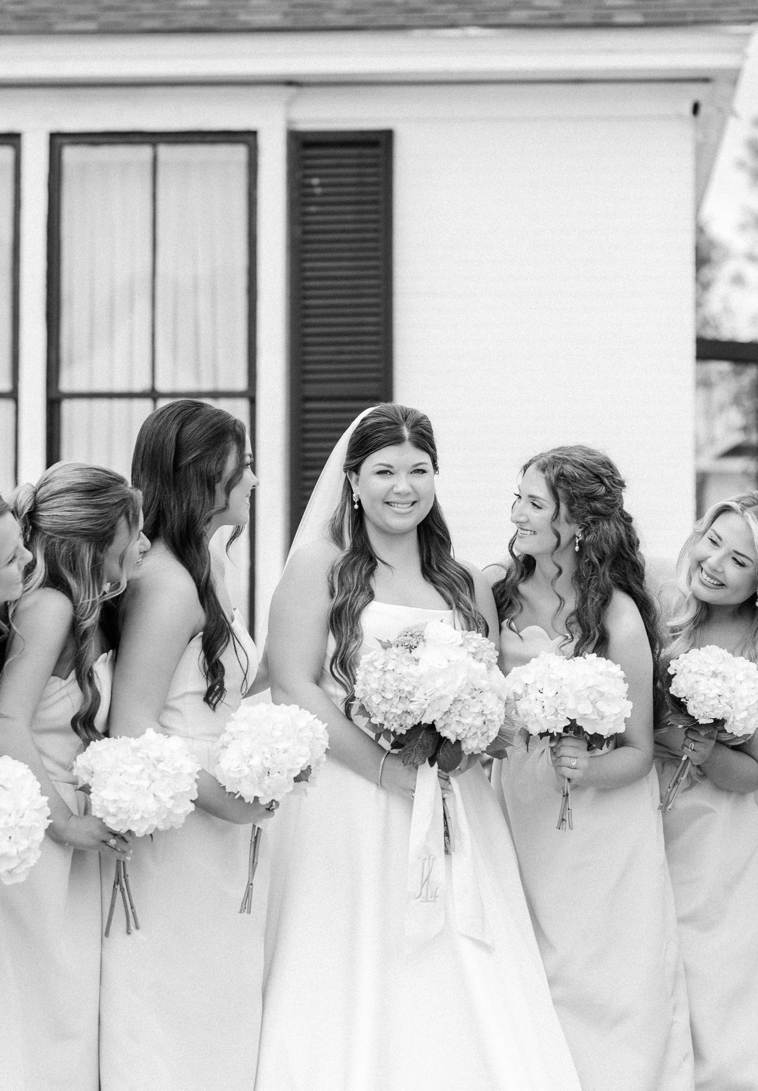 Black and white photo of a bride and bridesmaids holding white floral bouquets, smiling together outdoors.