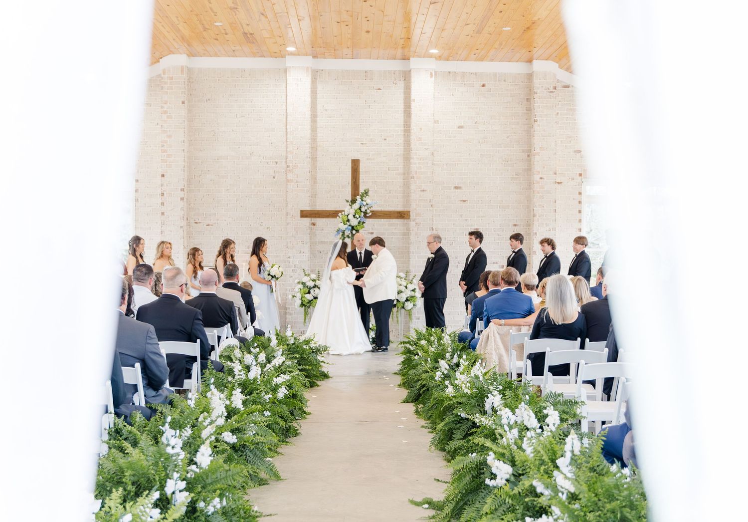 Elegant white chapel wedding ceremony with greenery aisle decor, cross altar, and seated guests.