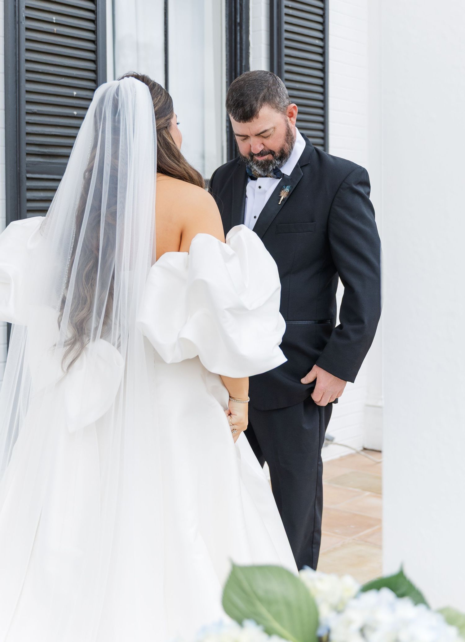 Bride in off-shoulder gown with veil shares first look moment with groom in black tuxedo outdoors.