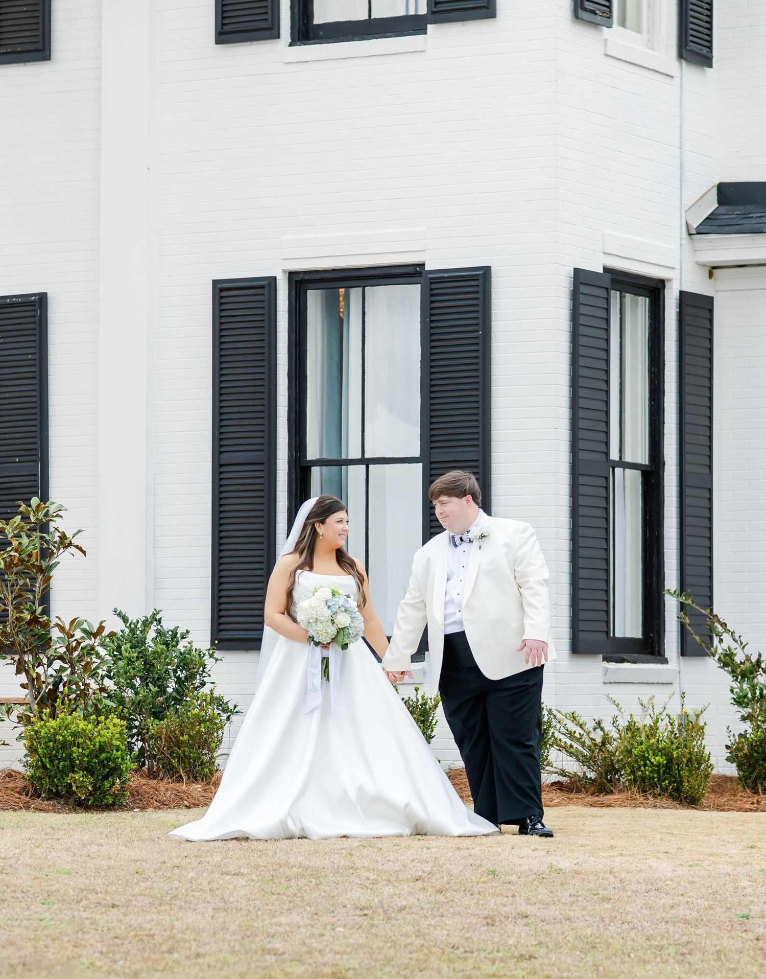 Bride and groom walk hand in hand outside a white Southern estate with black shutters on their wedding day.