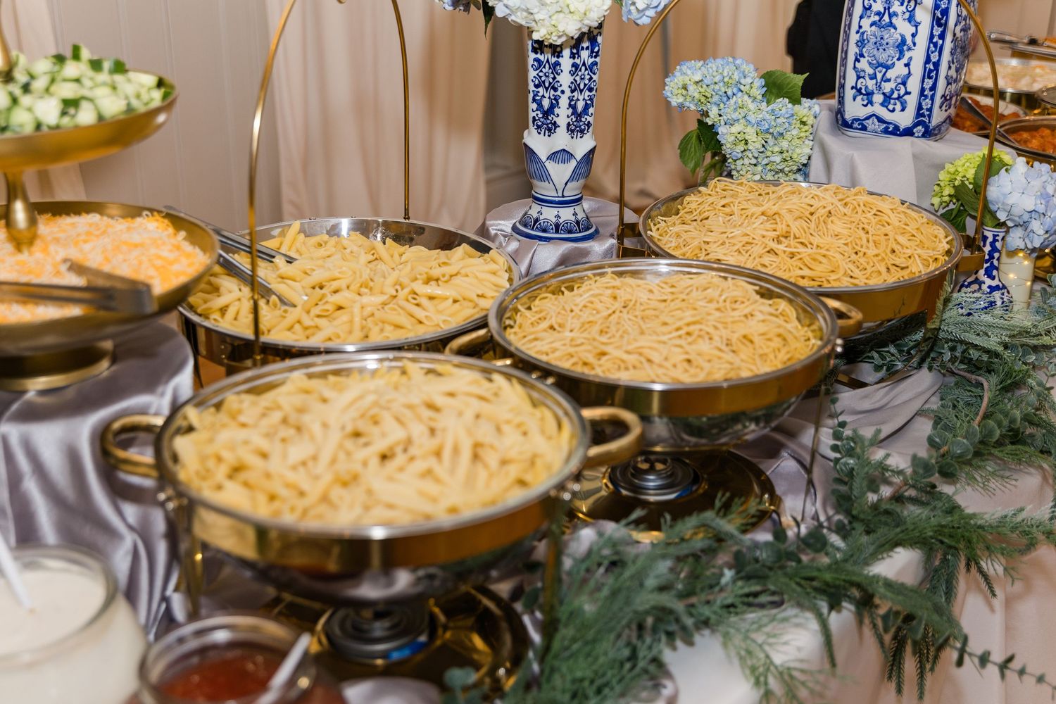 Elegant wedding reception buffet featuring multiple chafing dishes filled with pasta and fresh herb garnishes.