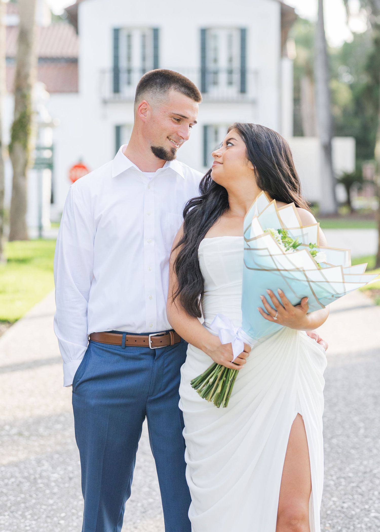 Smiling couple sharing a glance outdoors, woman in white dress holding floral bouquet, man in white shirt.