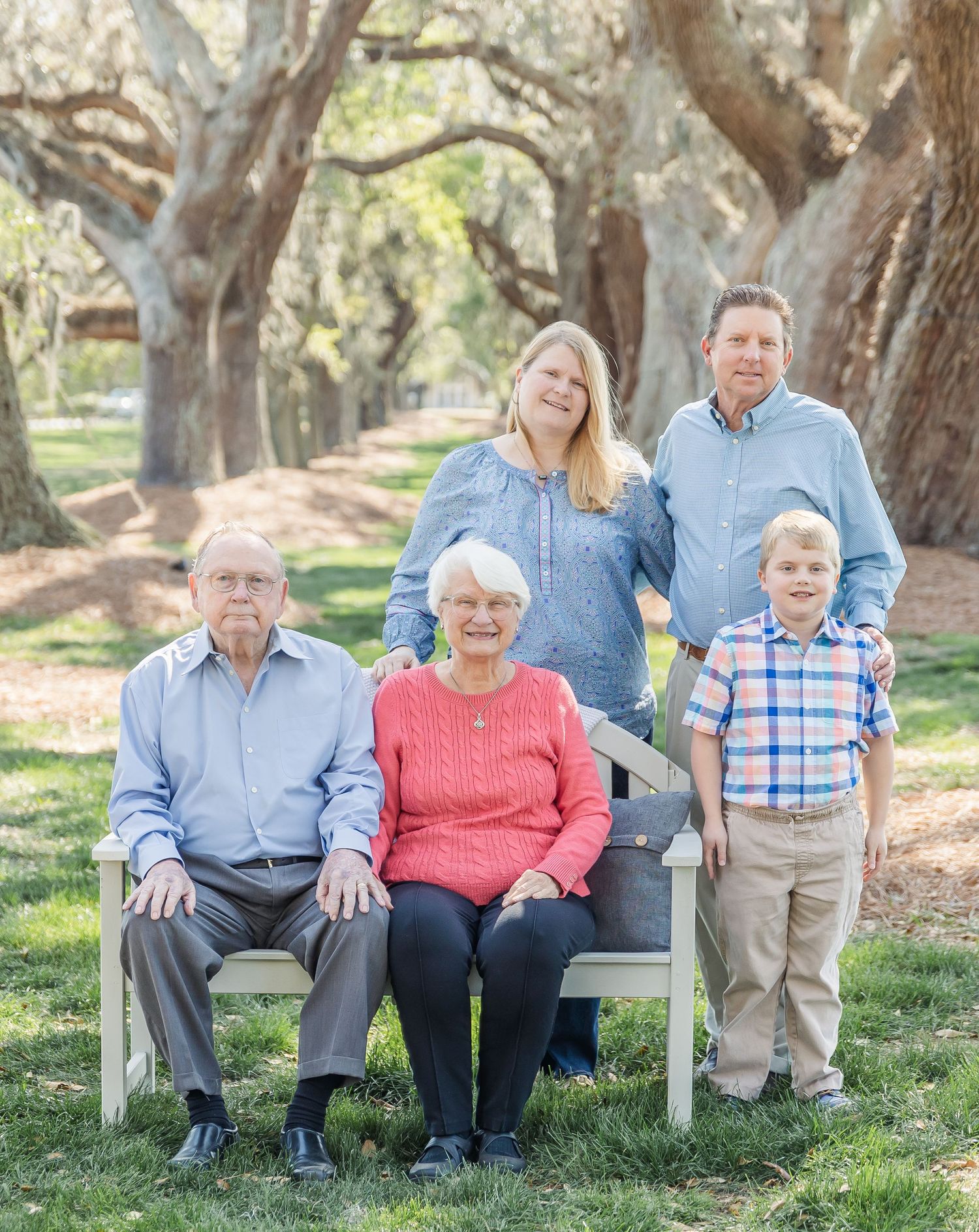 Multi-generational family poses together on a white bench surrounded by oak trees.