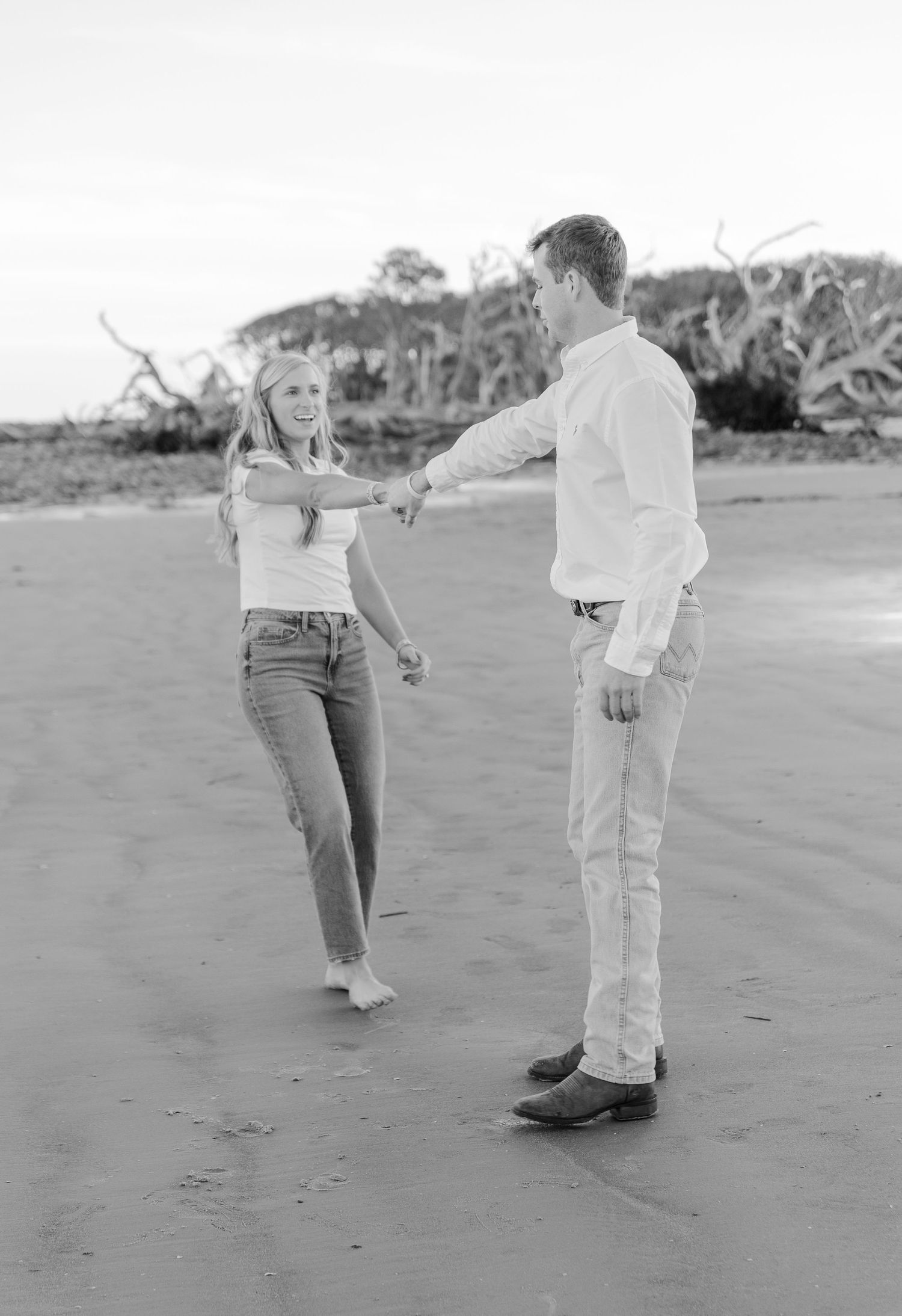 Black and white photo of a couple dancing on a beach with driftwood in the background.