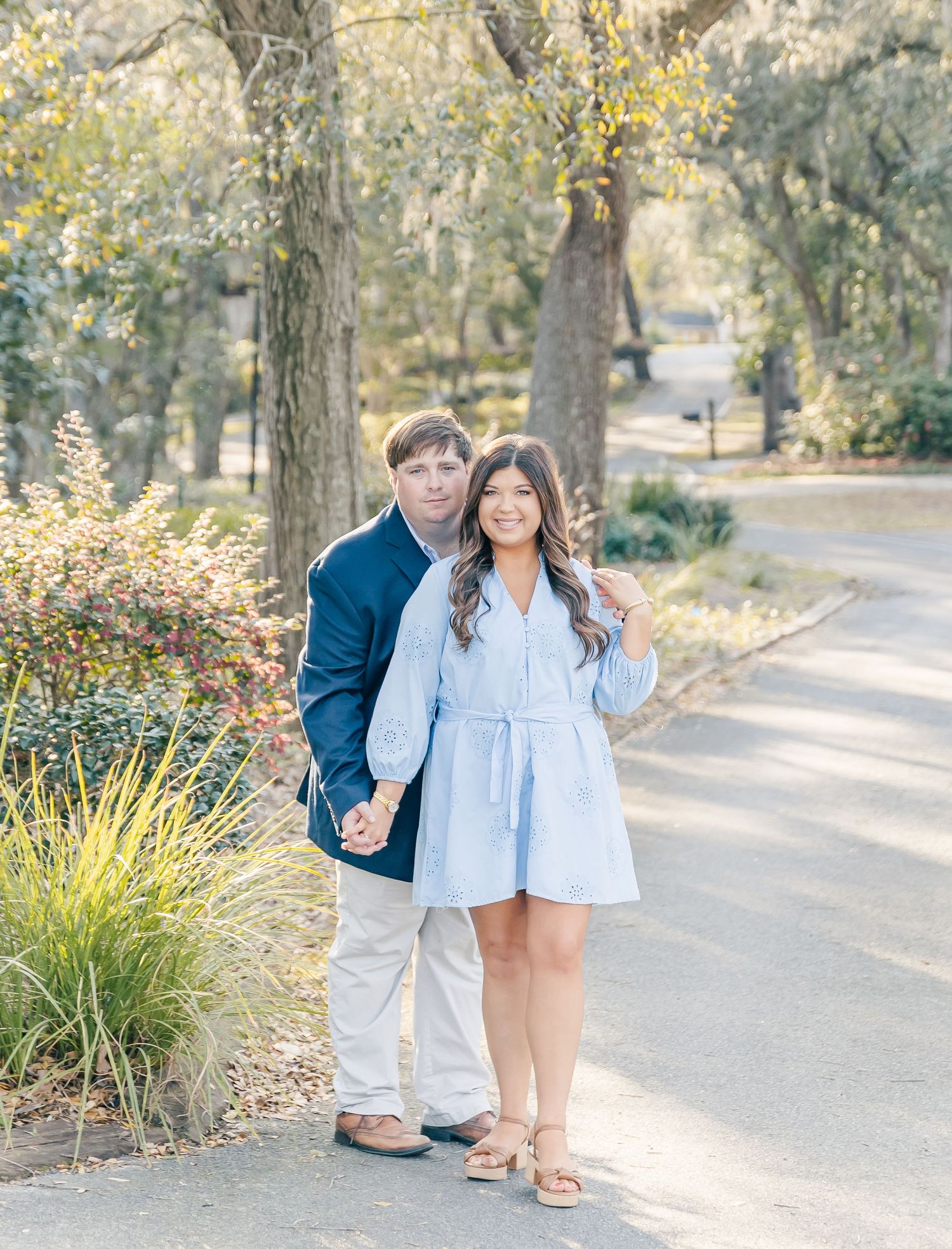 A couple poses together on a tree-lined path in casual spring attire.