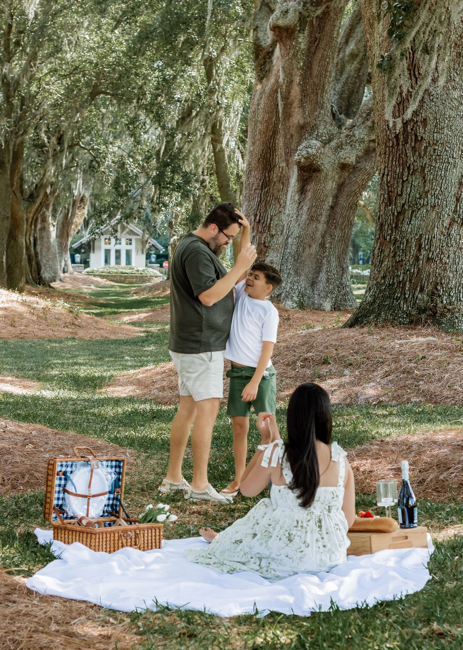 A family enjoys a picnic under large oak trees with a wicker basket on a white blanket.