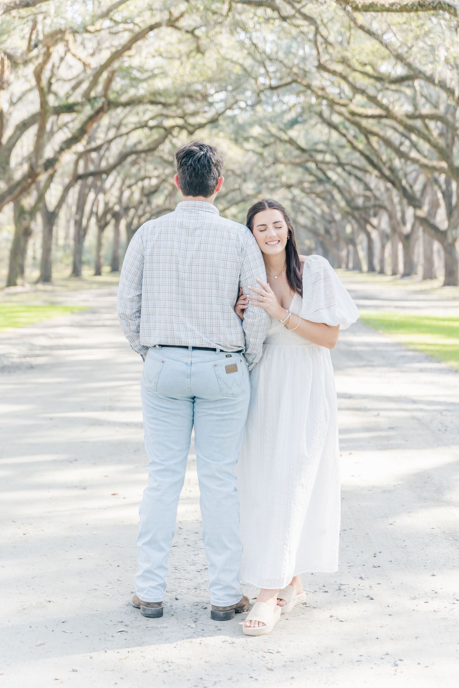 A romantic couple embraces on a path beneath an archway of oak trees.