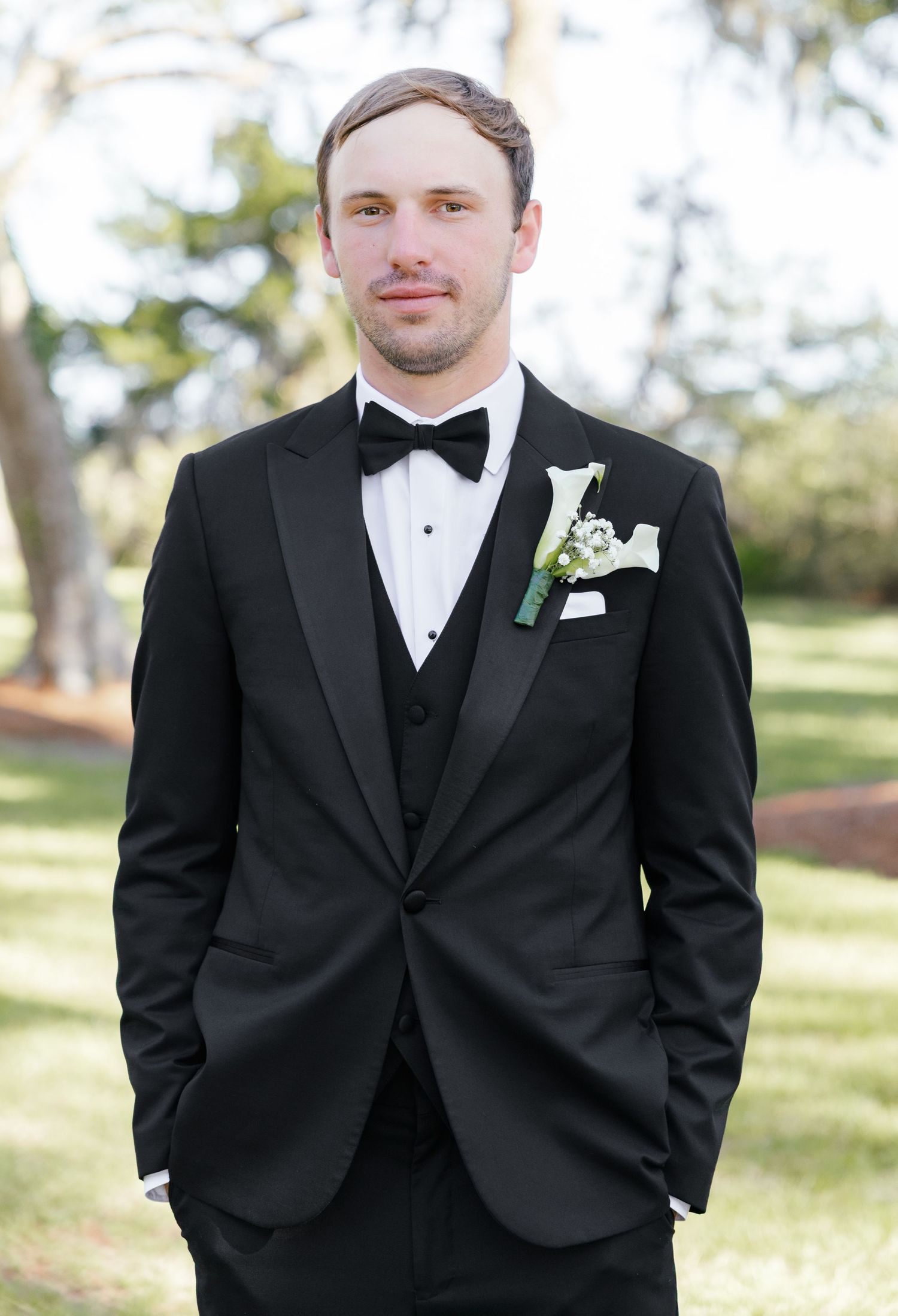 Groom in black tuxedo with bow tie and white floral boutonniere standing outdoors at wedding ceremony.