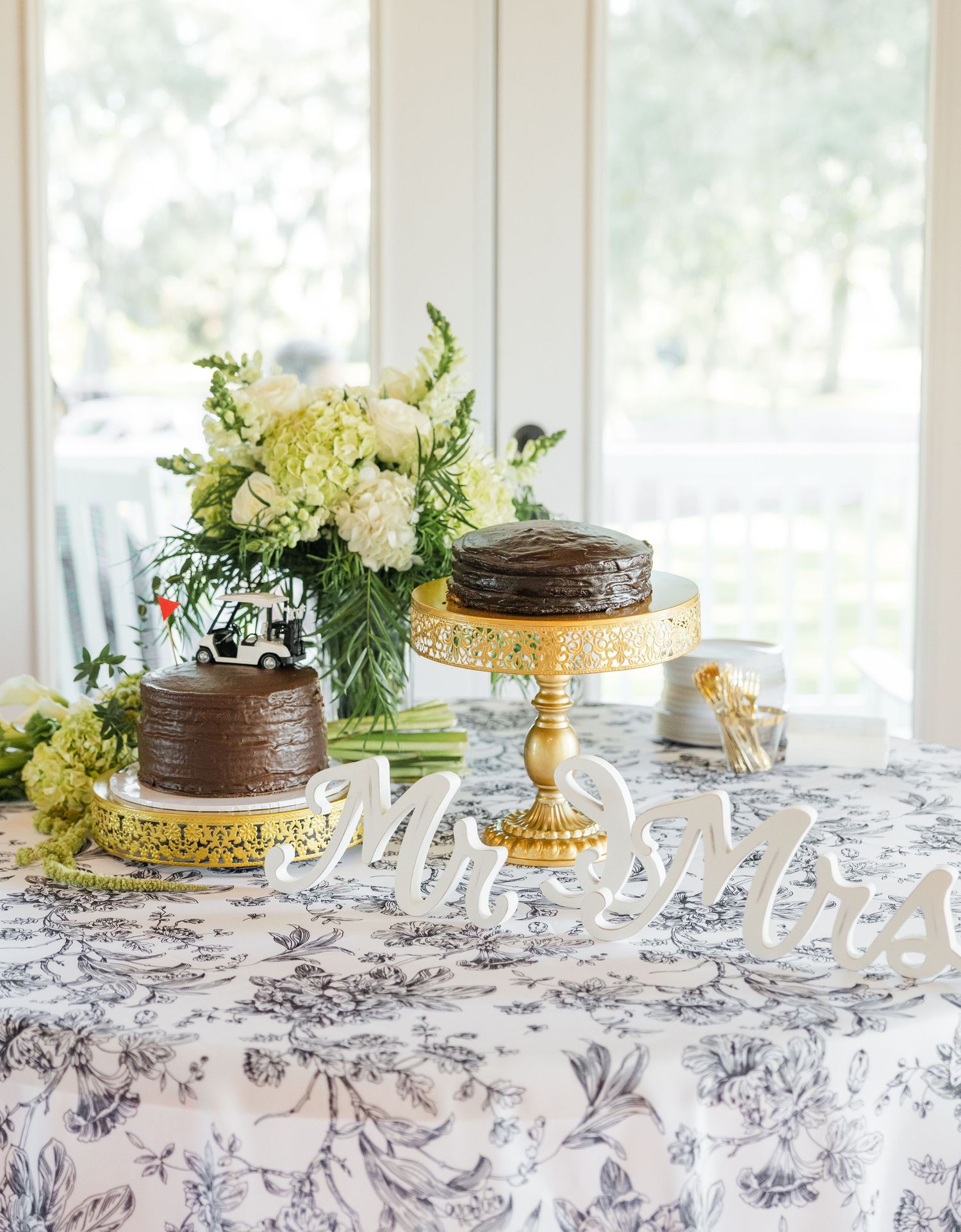 Wedding dessert table with chocolate cakes on gold stands, white floral arrangement, and Mr. & Mrs. signs on blue toile linen.