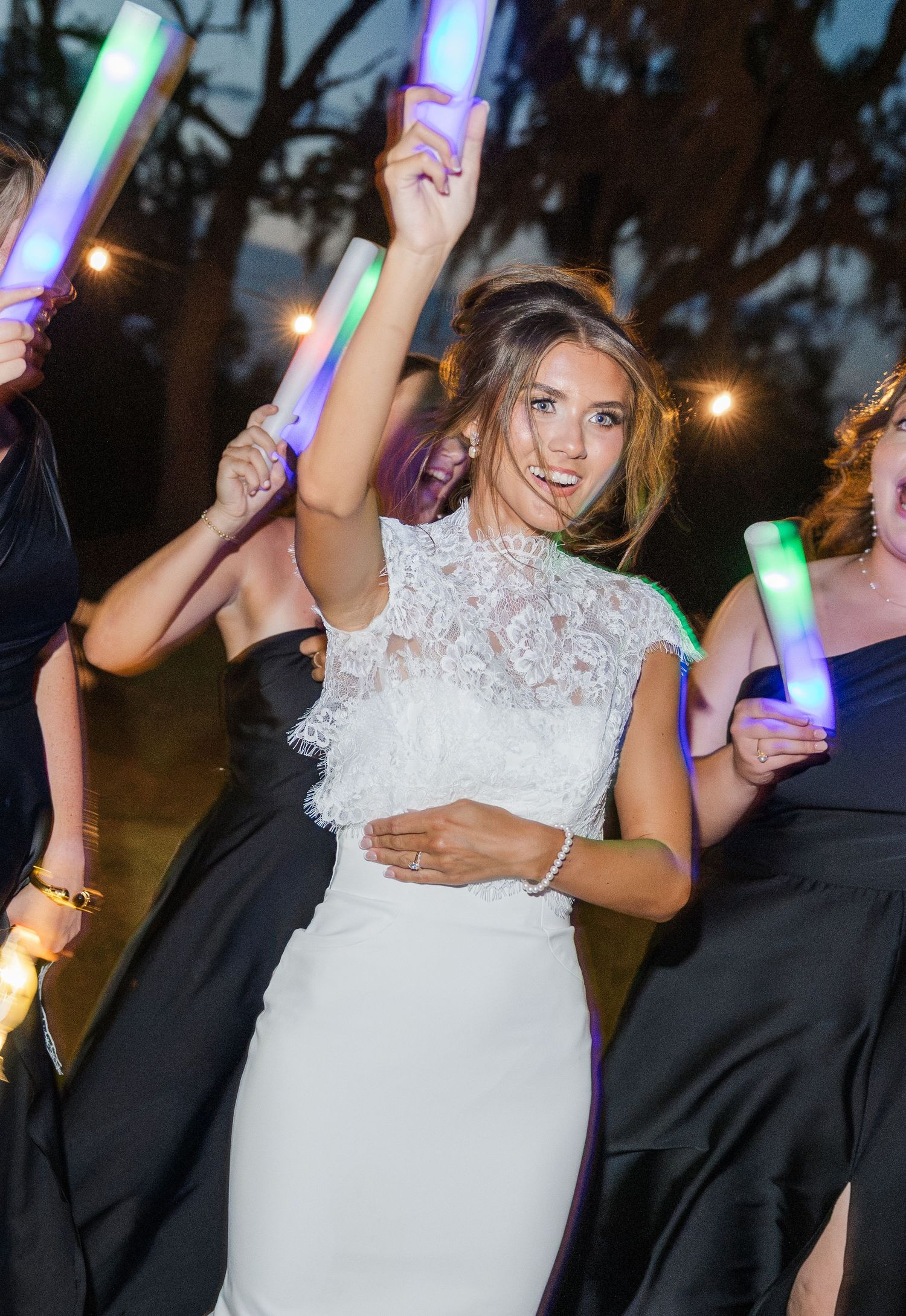 Joyful bride in white lace dress dancing with bridesmaids holding colorful LED glow sticks outdoors.