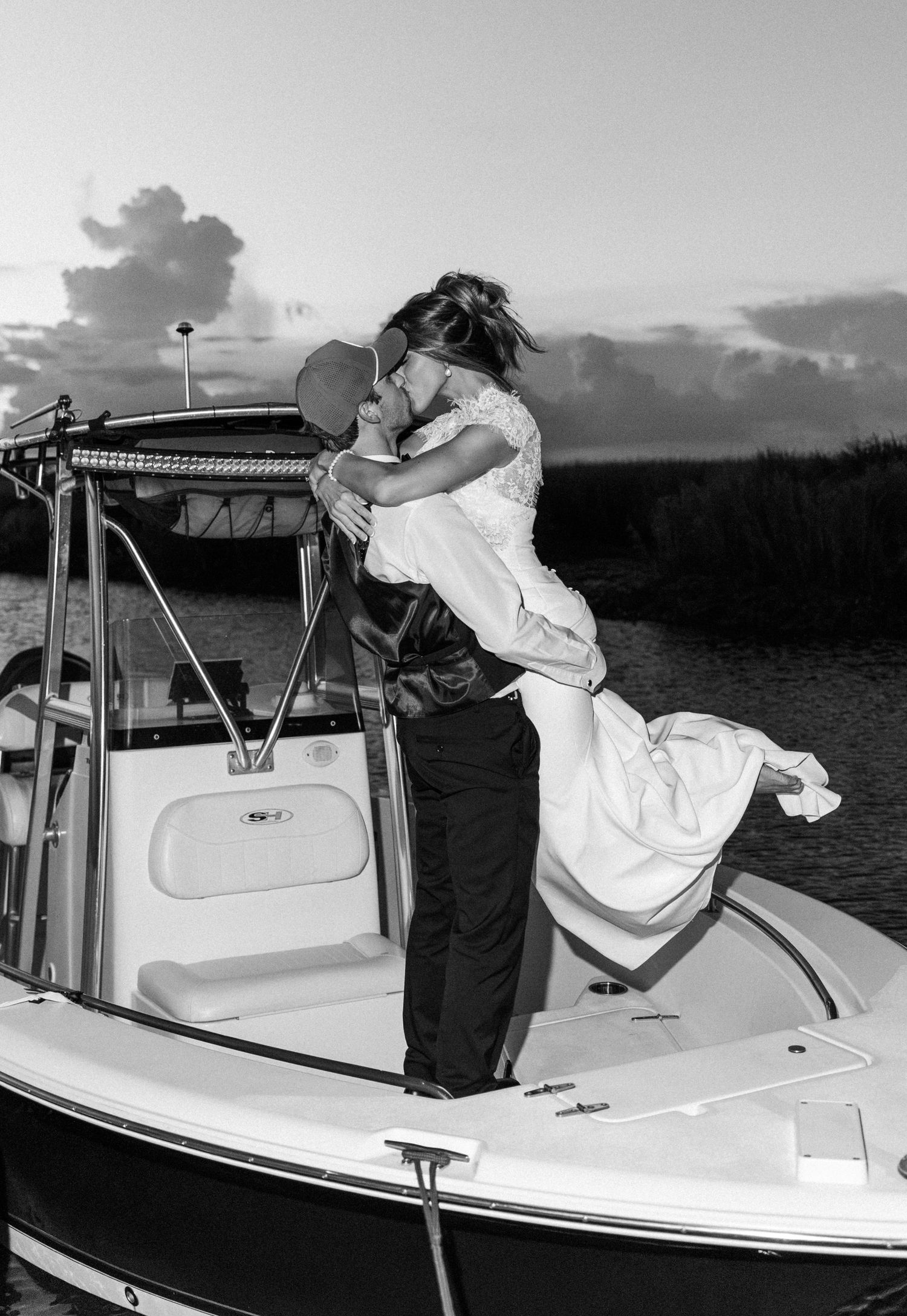 Black and white photo of groom lifting bride on a boat at sunset, sharing a romantic kiss on their wedding day.