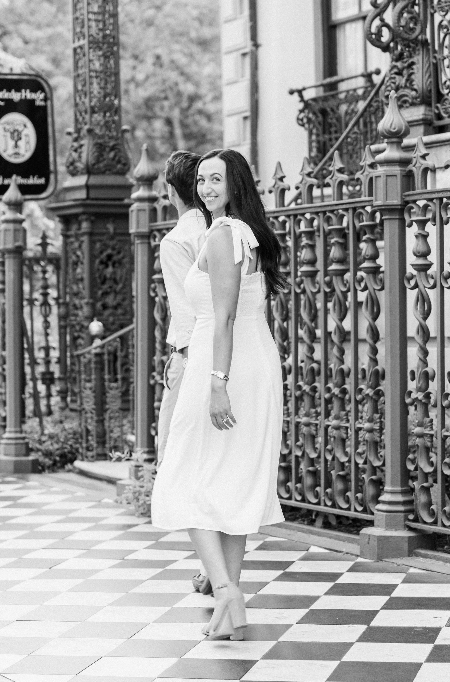 Black and white photo of woman in white dress looking back while standing by ornate iron gates.