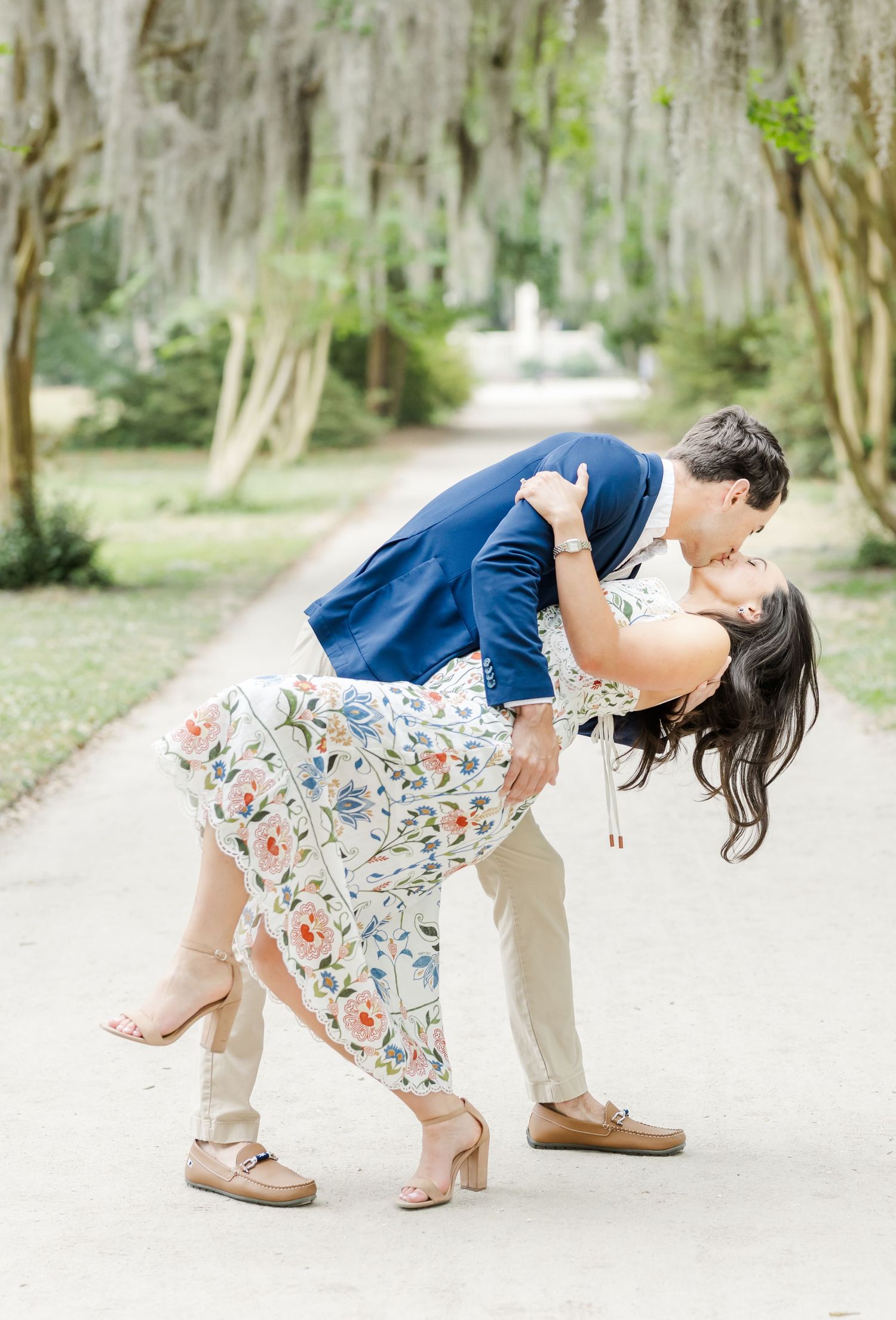 Couple sharing a romantic dip kiss on a tree-lined path, woman in floral dress, man in navy blazer.