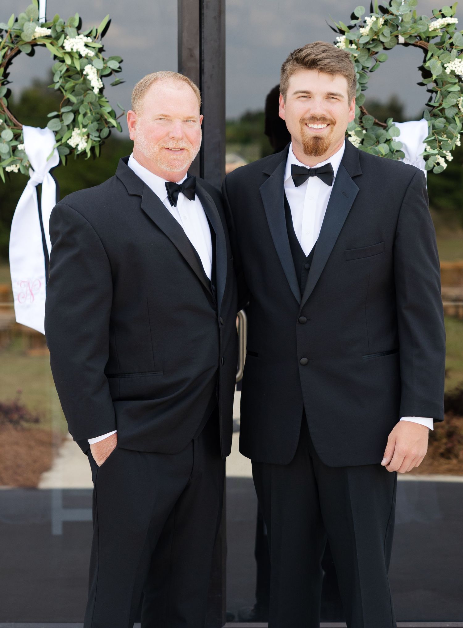 Two groomsmen in black tuxedos with bow ties smile in front of greenery-adorned wedding venue doors.