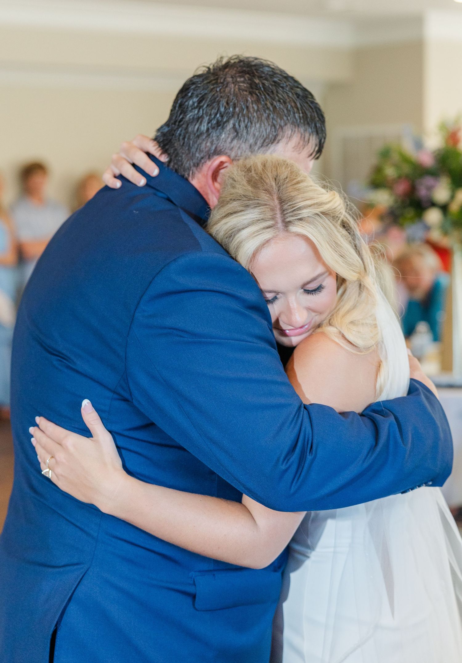 Emotional bride in white gown shares a heartfelt hug with a man in a navy blue suit at reception.