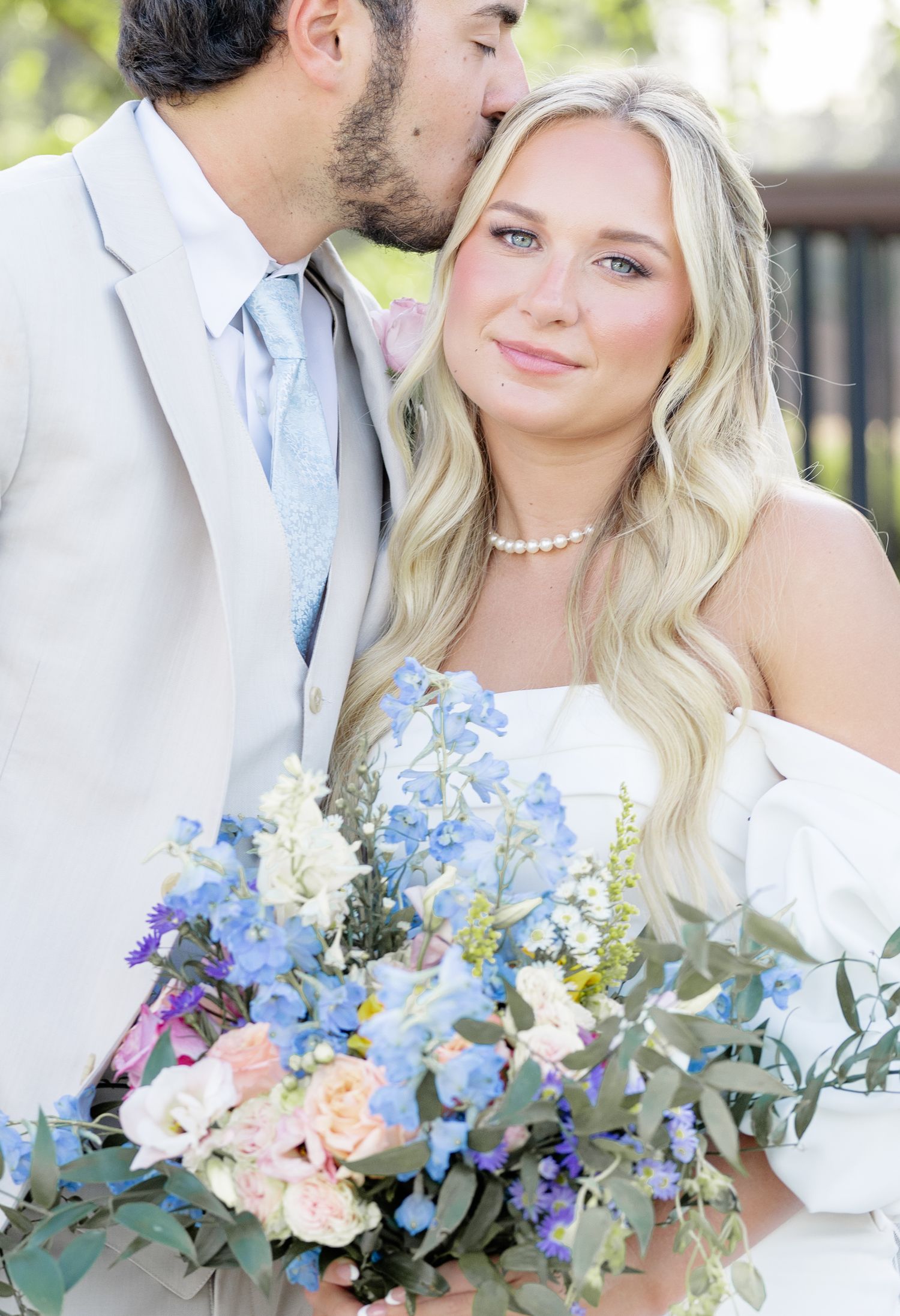 Groom in tan suit kissing bride on cheek as she holds blue and white floral wedding bouquet outdoors.