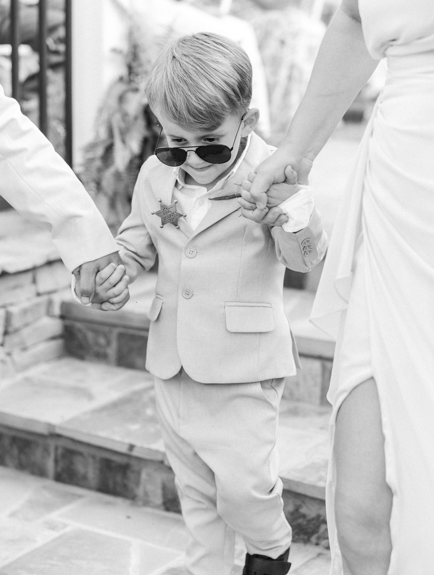 Adorable young ring bearer in tan suit and sunglasses holding hands walking down steps.