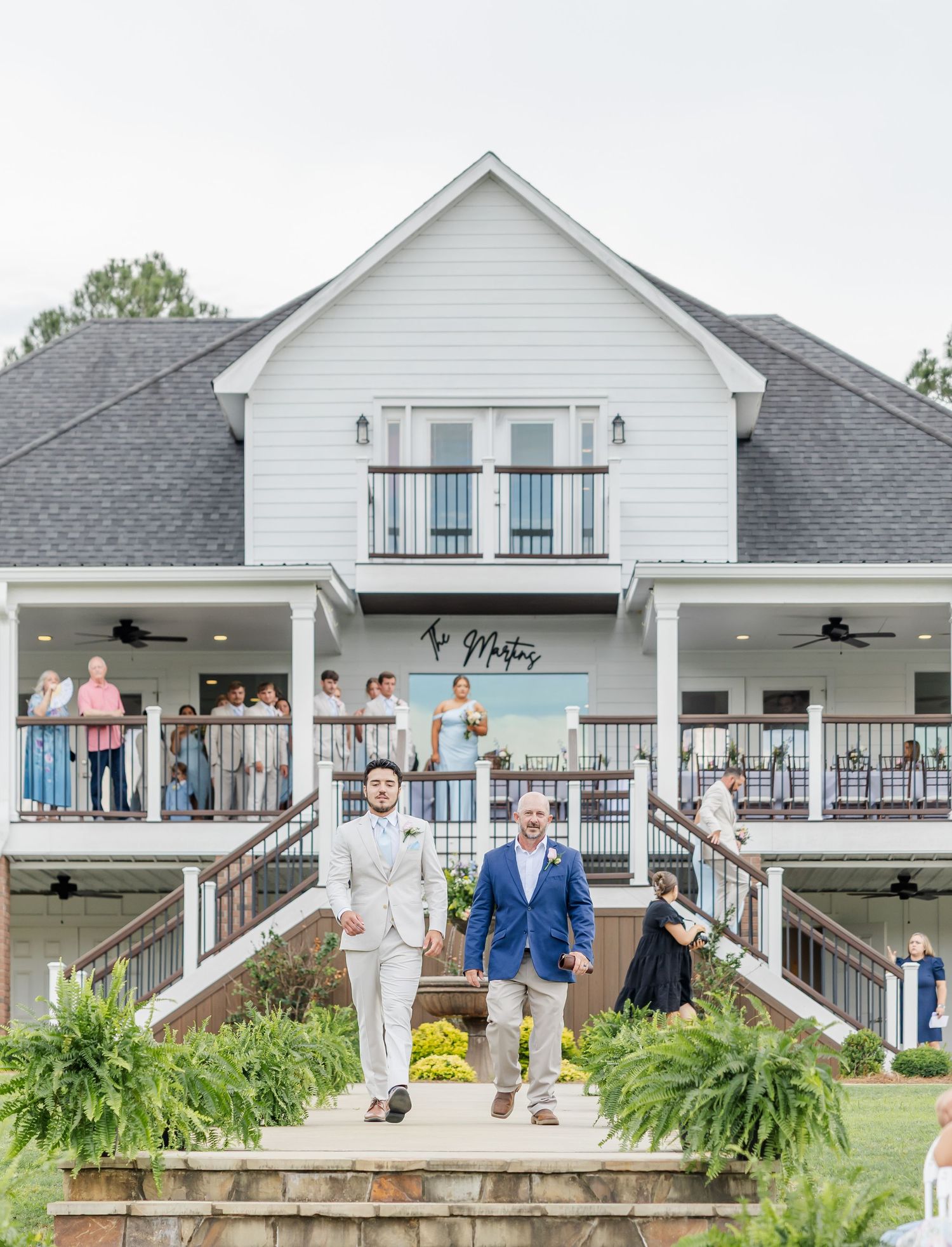 Two grooms walk down steps of a white farmhouse wedding venue with guests watching from balcony.