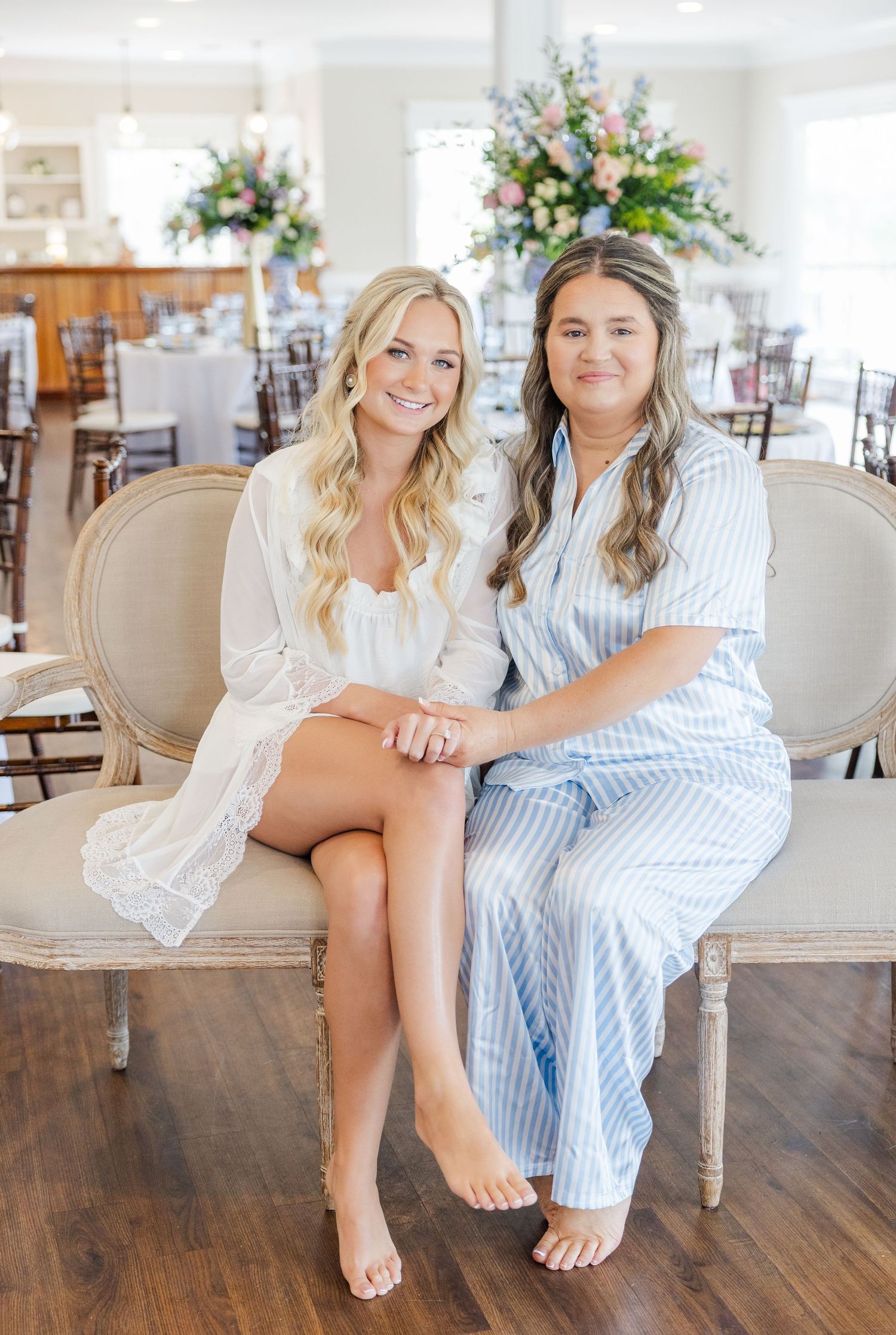 Bride in white lace robe and woman in blue striped pajamas sitting together on vintage settee.