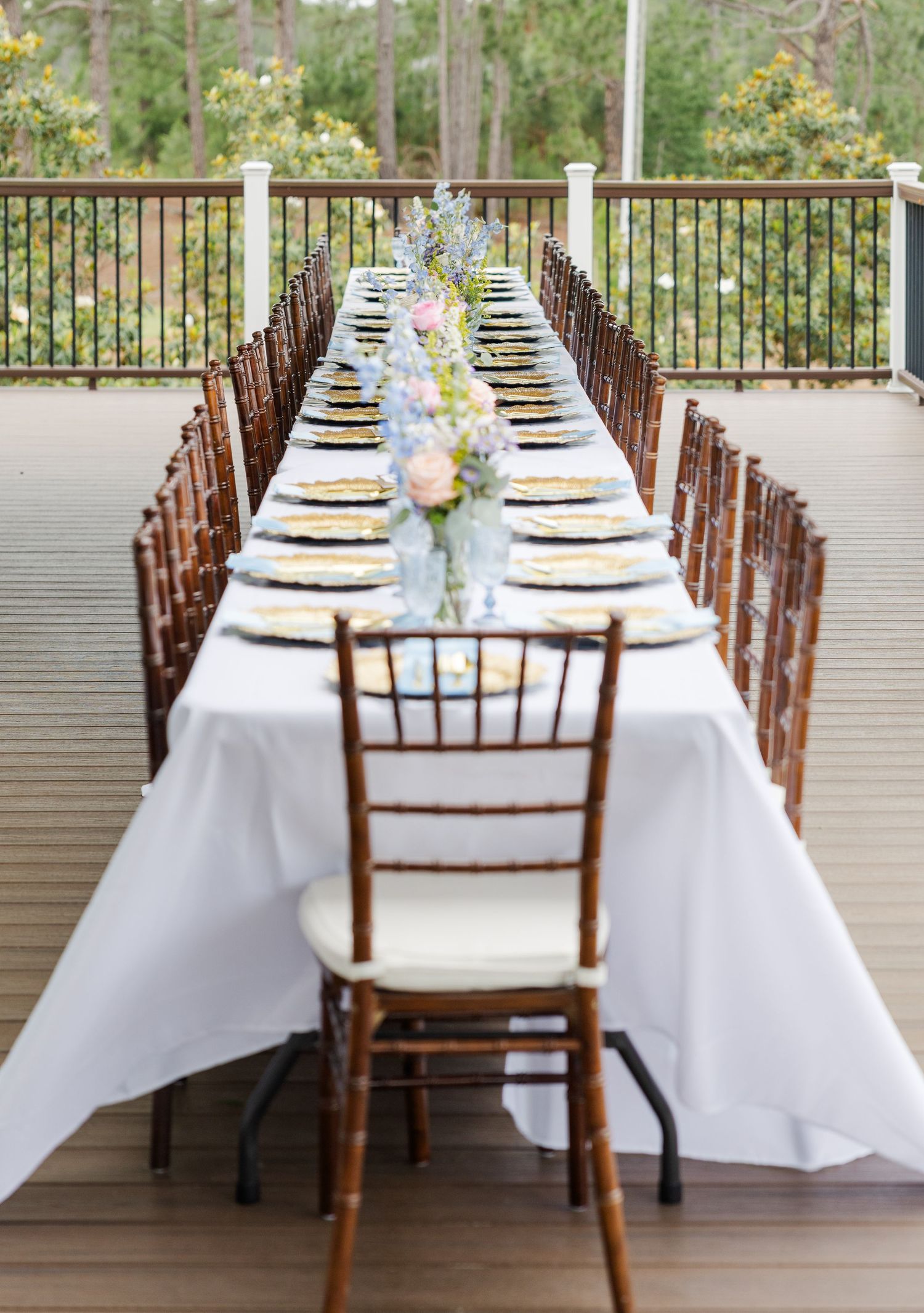 Long outdoor reception table with white linen, gold chargers, and pastel floral centerpieces on wooden deck.