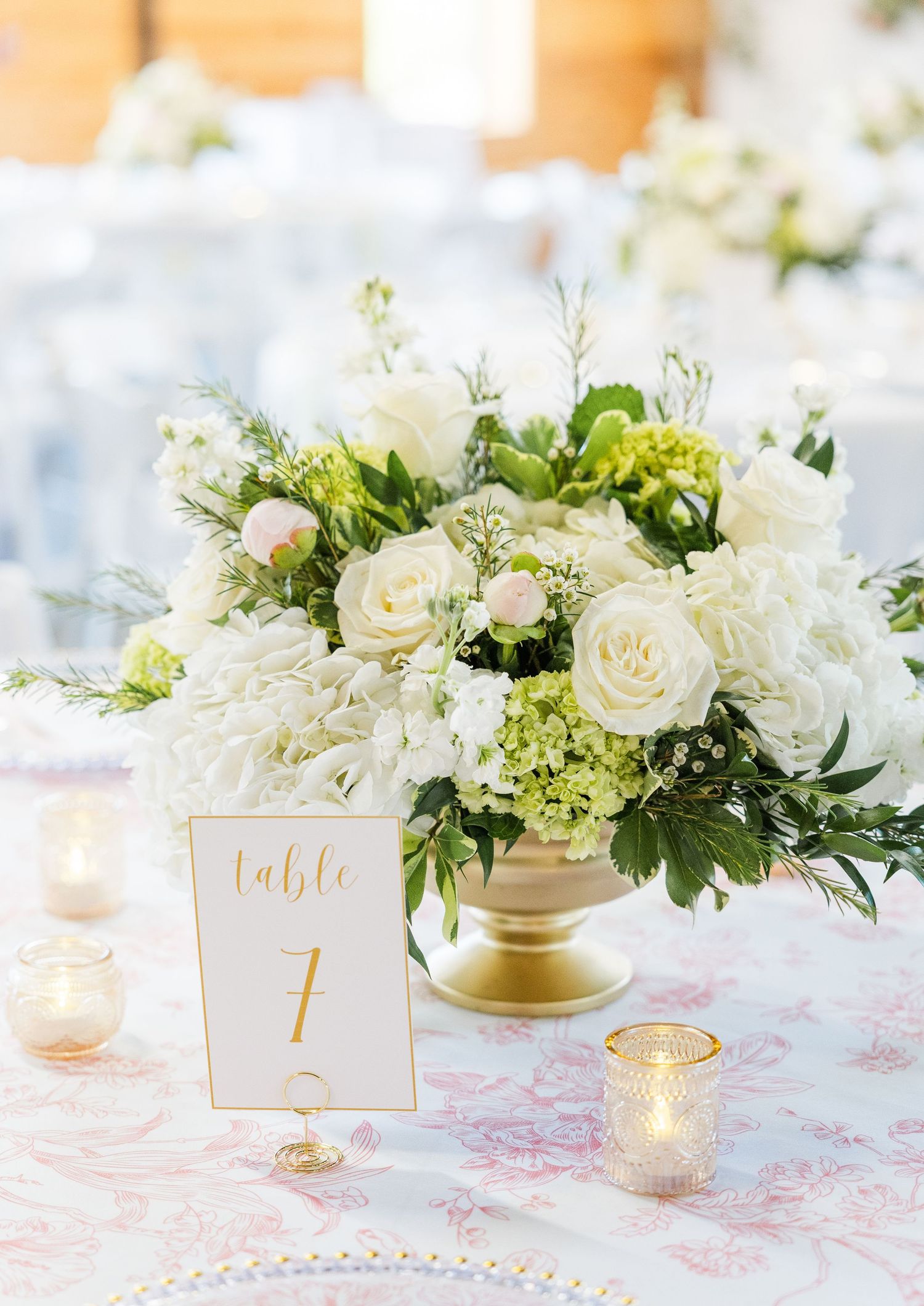 Elegant white rose and hydrangea wedding centerpiece in gold urn vase with Table 7 card and votive candles.