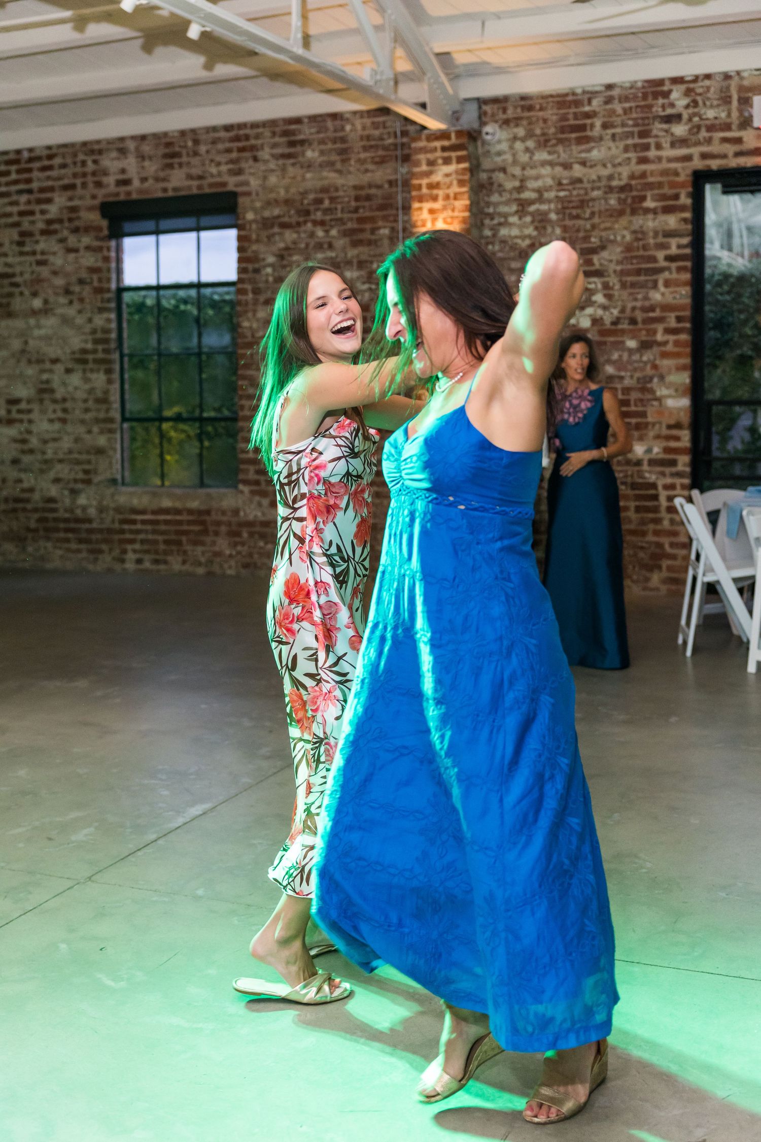 Two women in colorful dresses dance joyfully under green lights at wedding reception.
