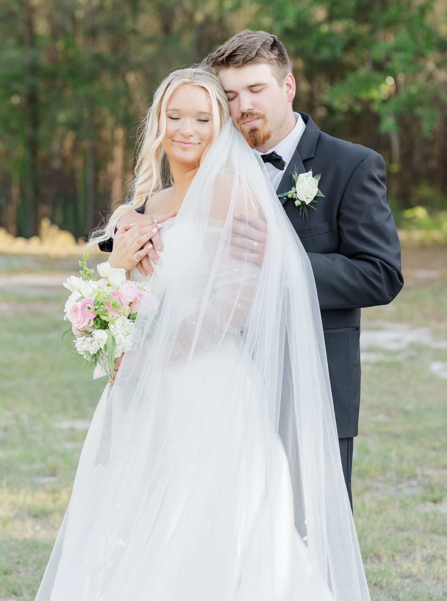 Bride in white gown with veil and groom in black tuxedo sharing a tender moment outdoors.