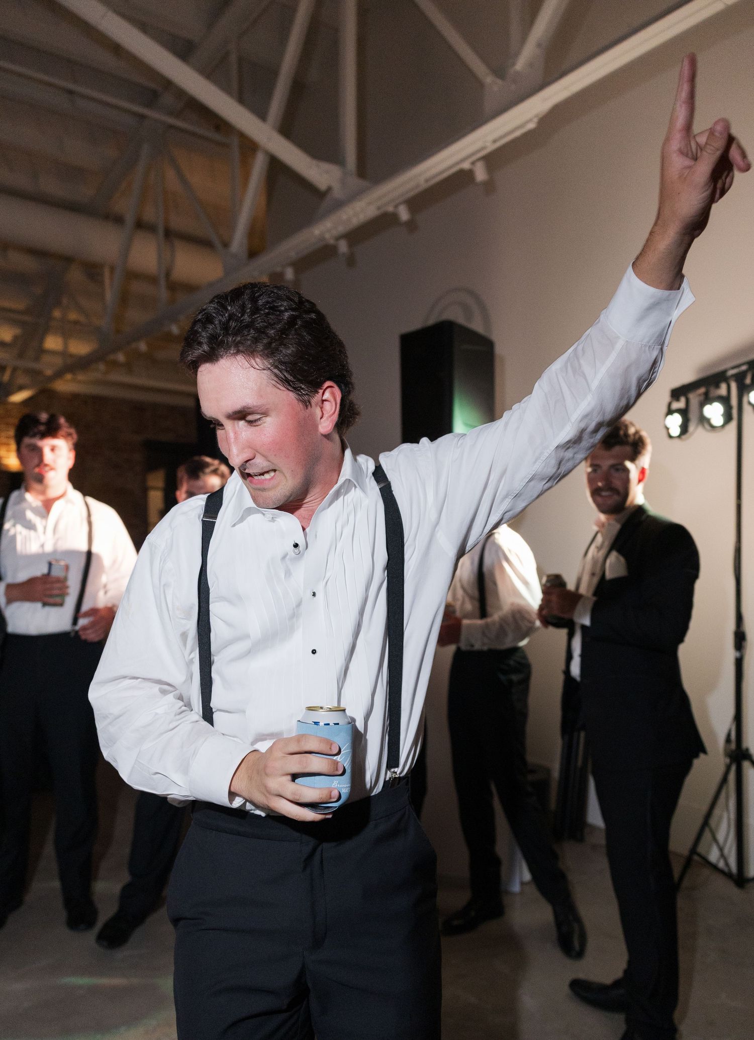 Man in white shirt and suspenders dances with finger pointed up holding drink at wedding.