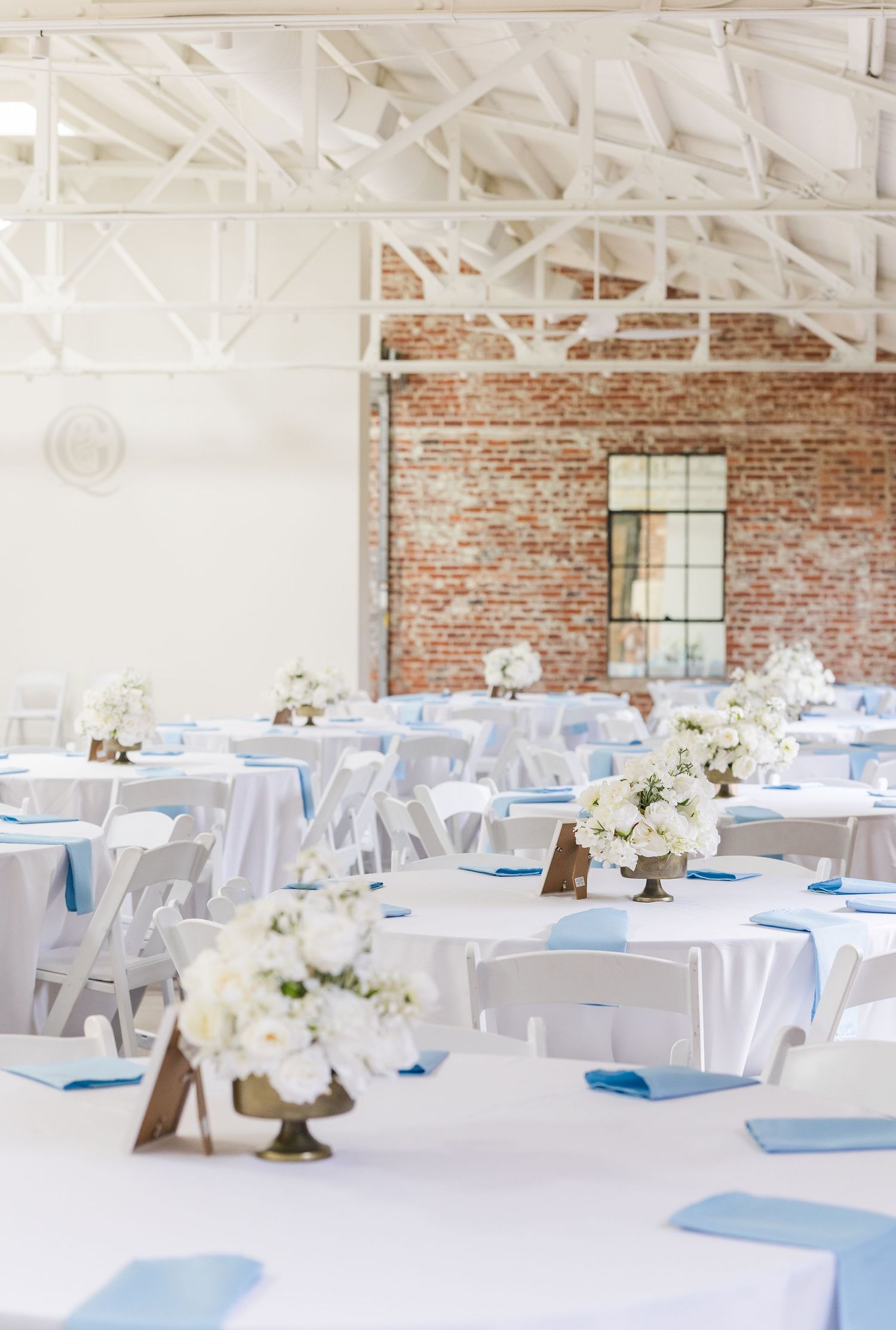 Elegant wedding reception hall with white tables, blue napkins, floral centerpieces, exposed brick walls, and white steel truss ceiling.
