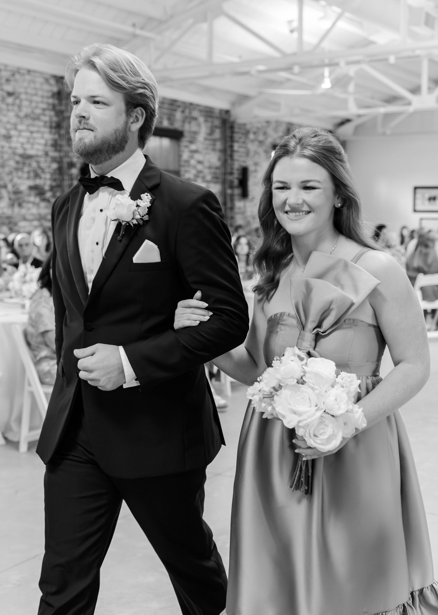 Black and white photo of groomsman escorting smiling bridesmaid down the aisle.