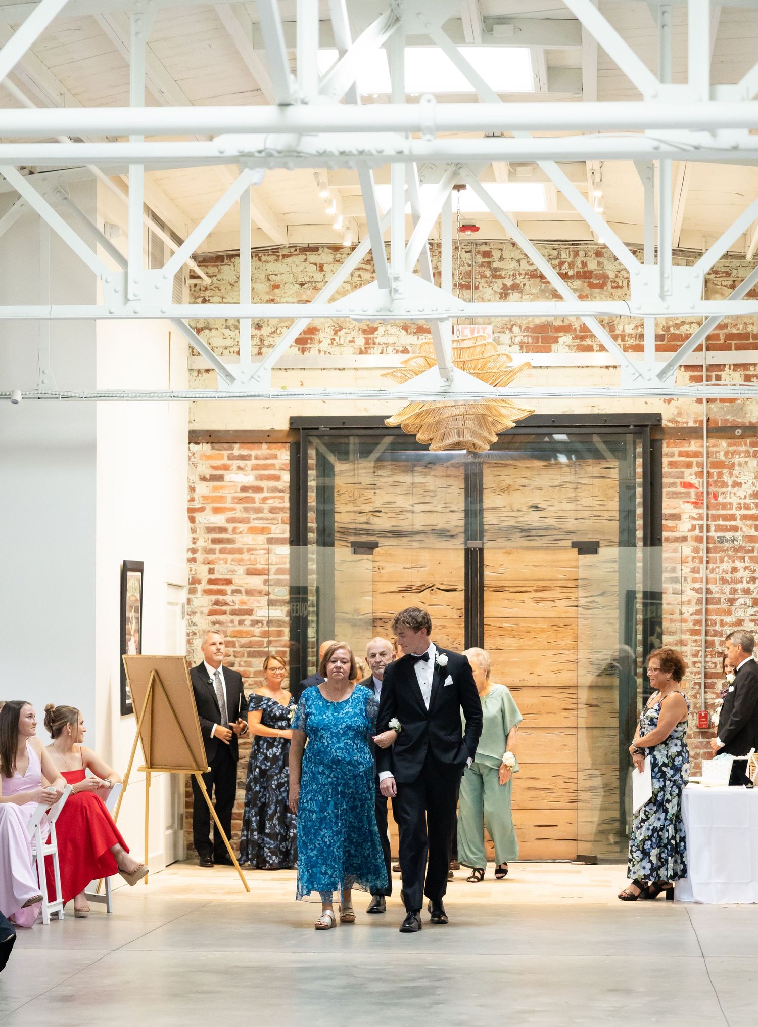 Groom escorts woman in blue floral dress down wedding ceremony aisle in industrial brick venue.