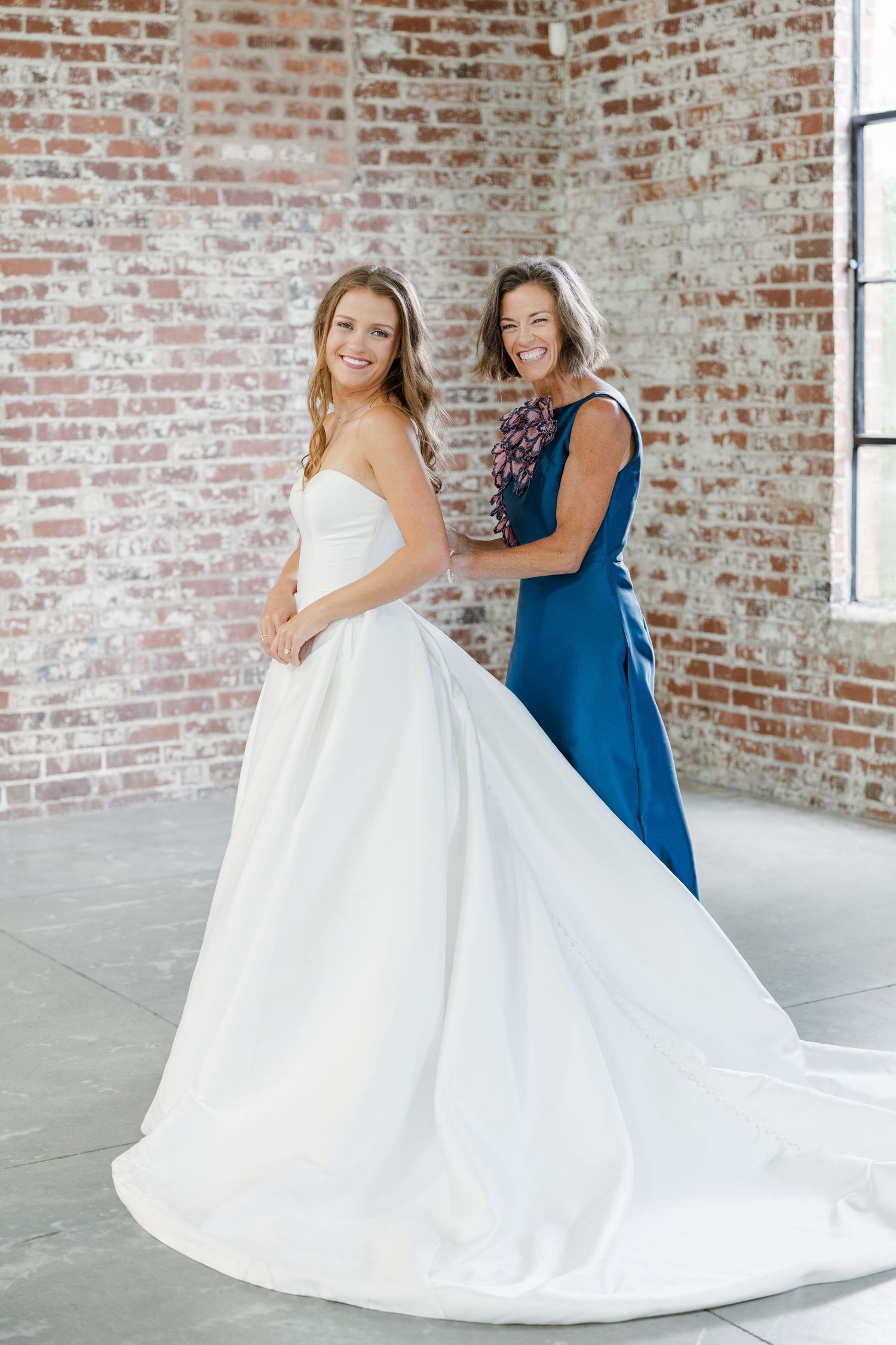 Laughing bride in white gown with woman in blue dress against exposed brick wall background.