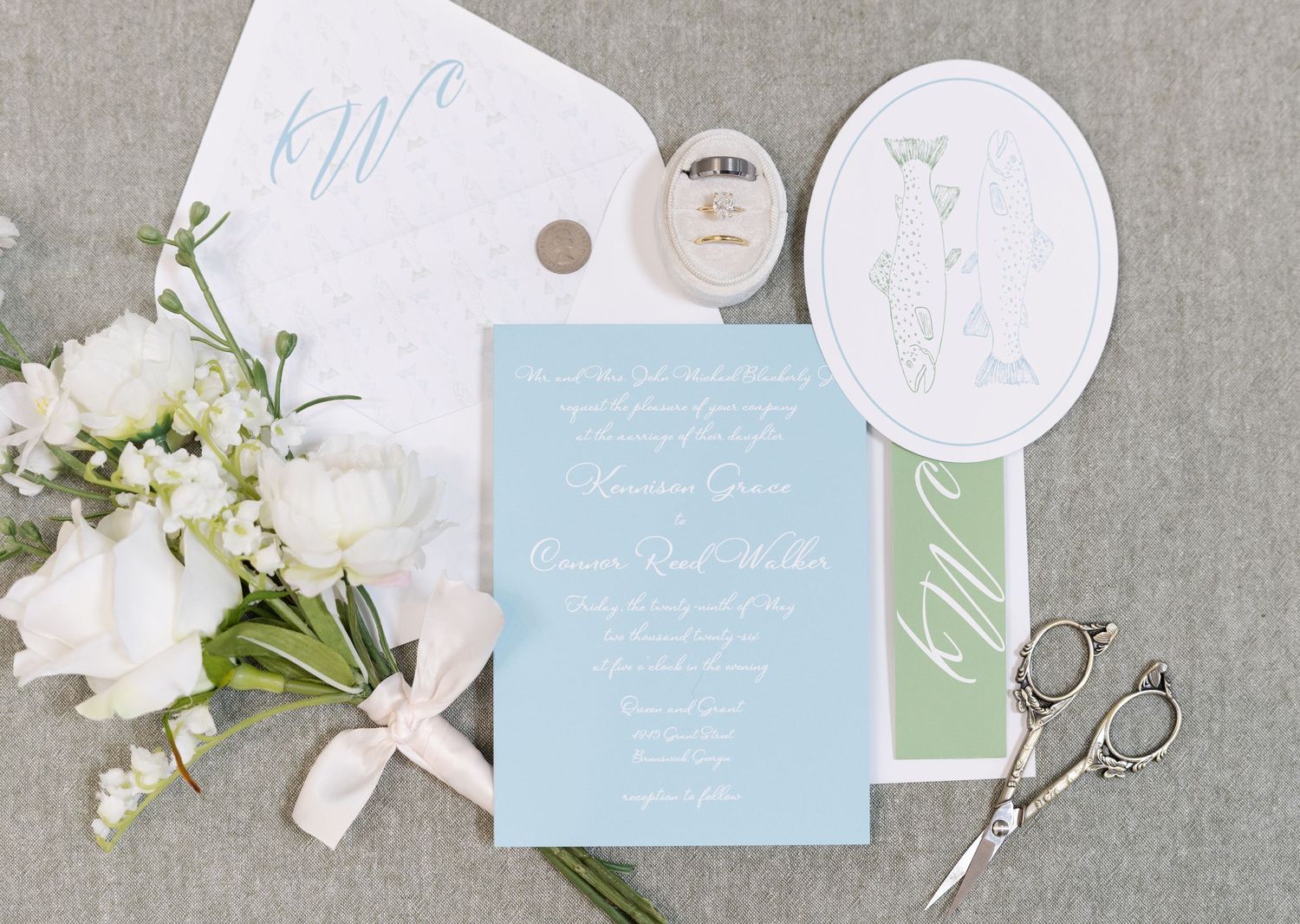 Light blue wedding invitation suite with floral bouquet, monogram, scissors, and oval illustration.