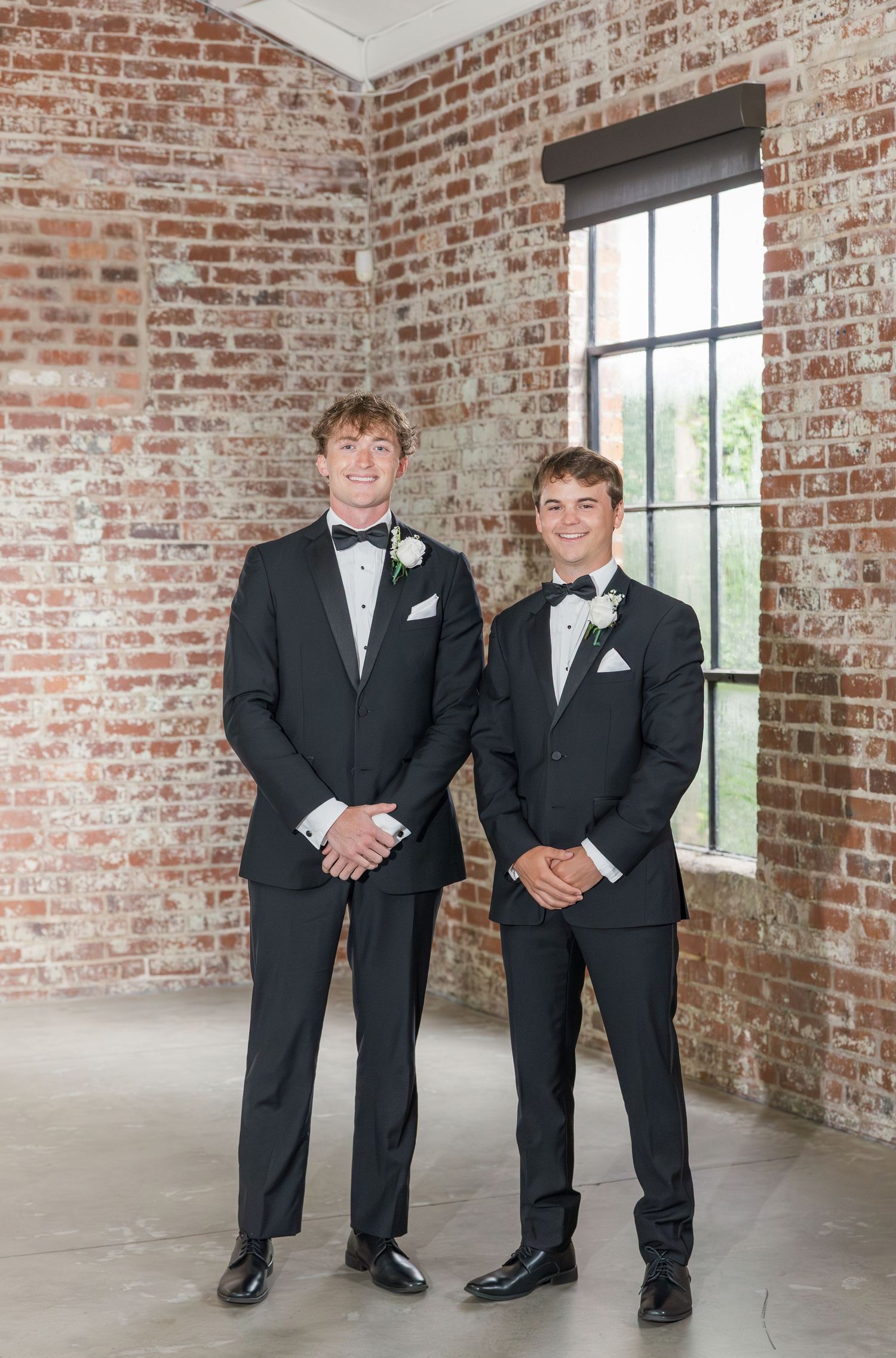 Two young men in black tuxedos with bow ties and boutonnieres pose in front of a rustic brick wall.