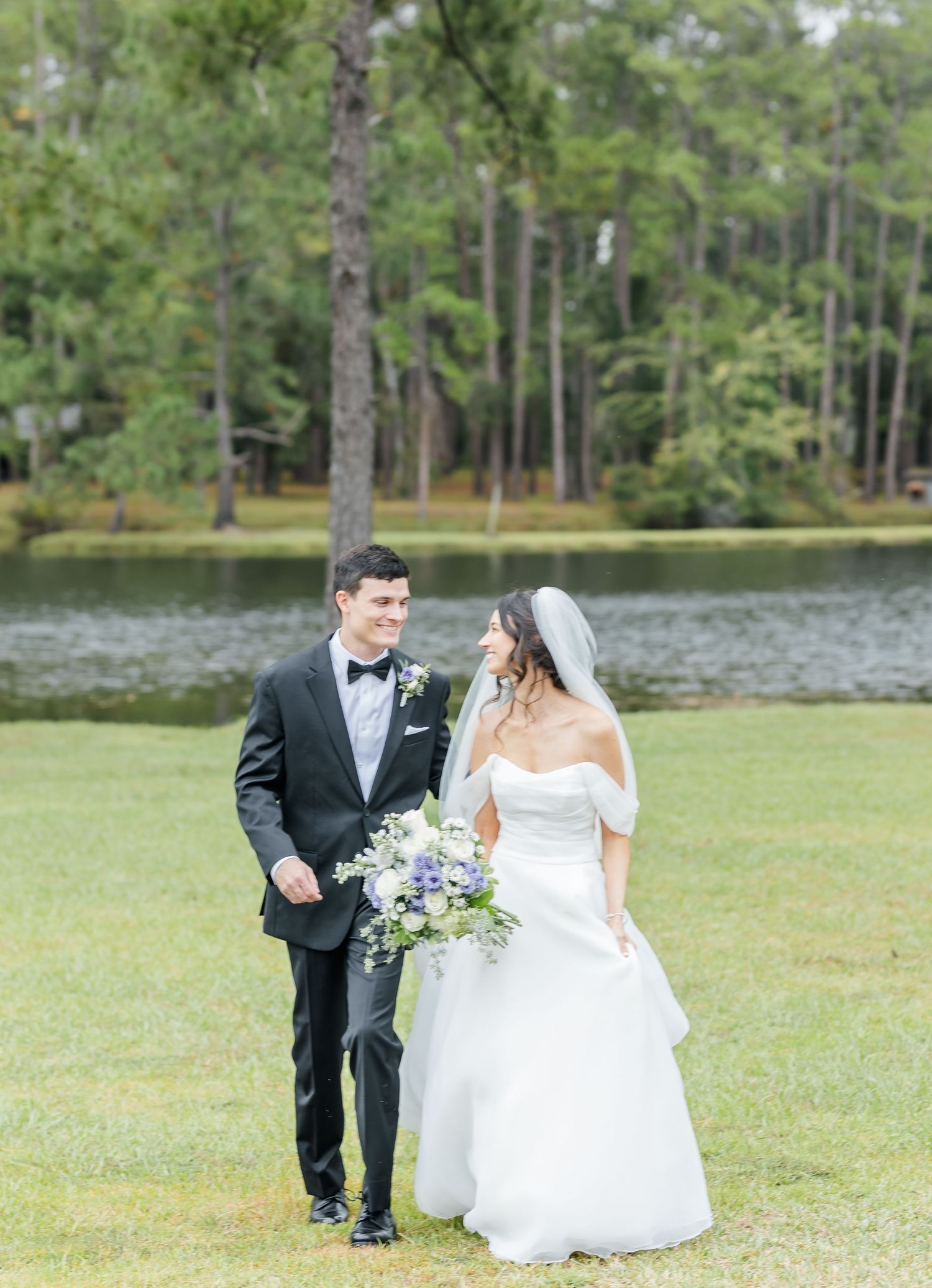 Newlyweds walk together on grass near a serene lake surrounded by pine trees.