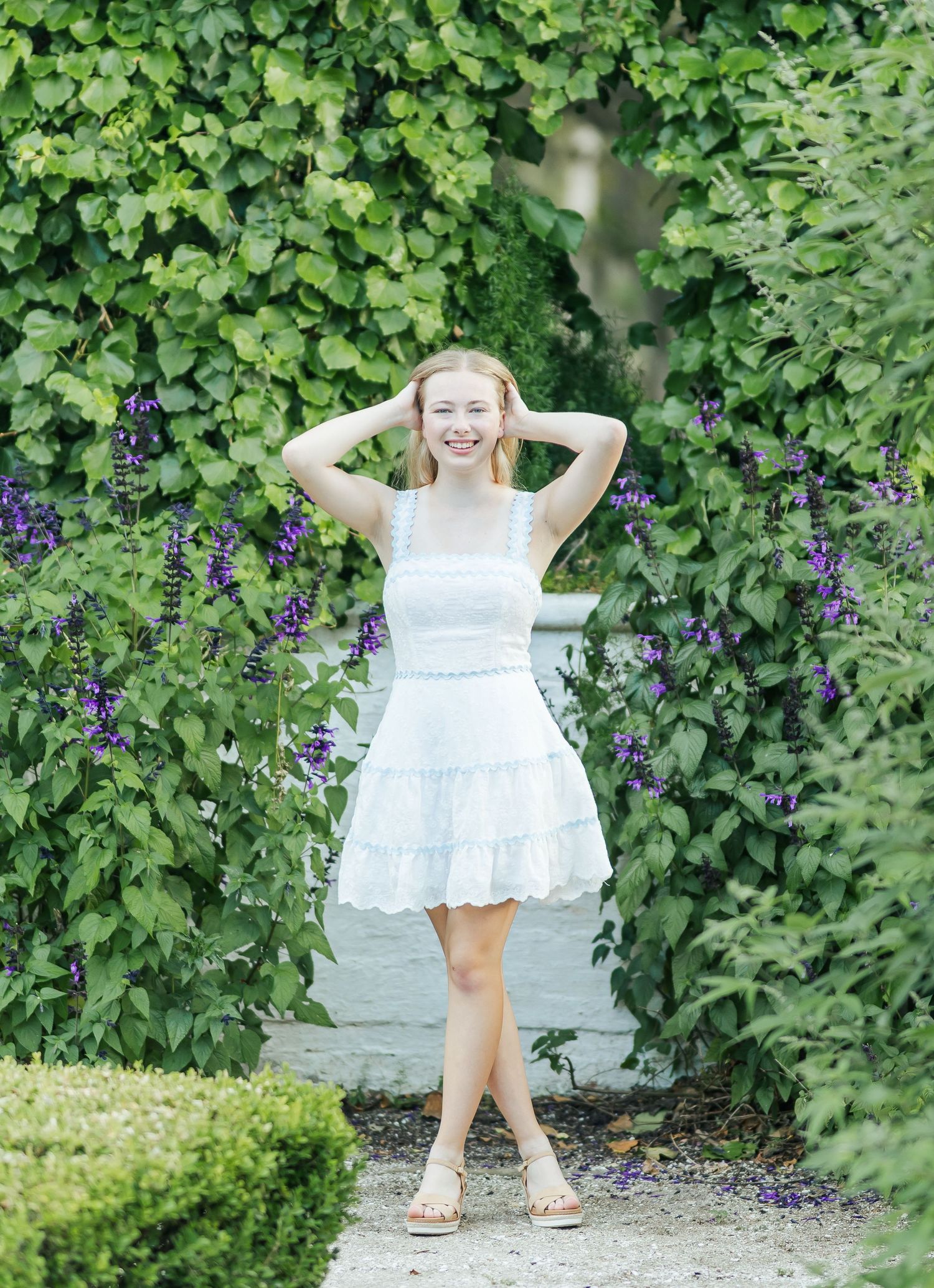 A person in a white lace dress poses playfully in a garden setting with purple flowers and greenery.