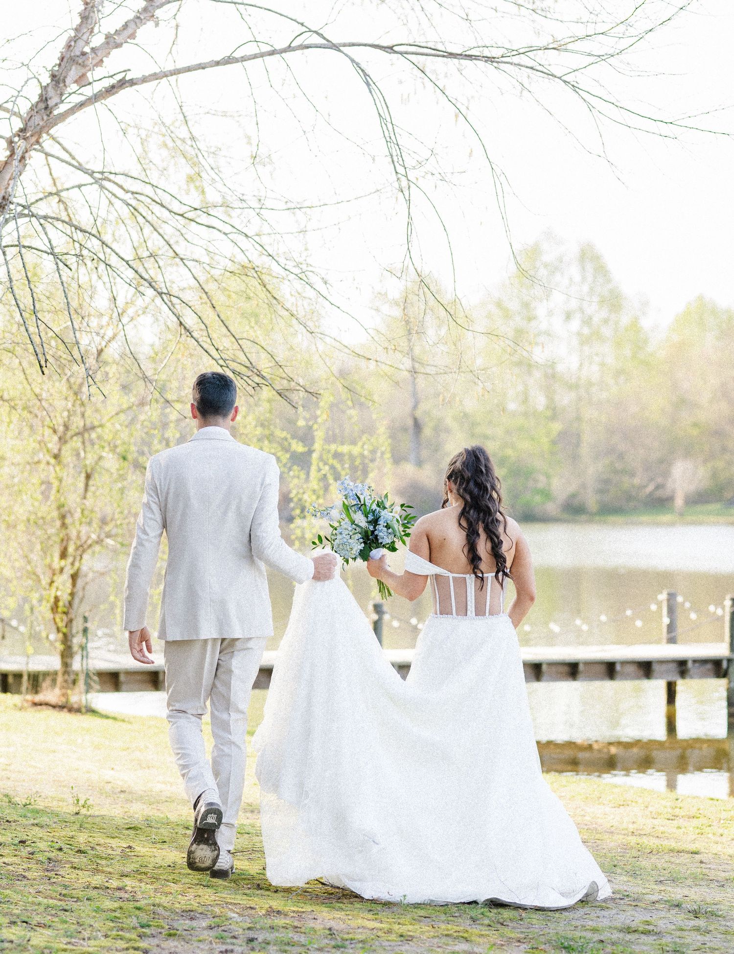 Bride and groom walking hand-in-hand toward lakeside dock, bride in backless lace gown holding blue bouquet.