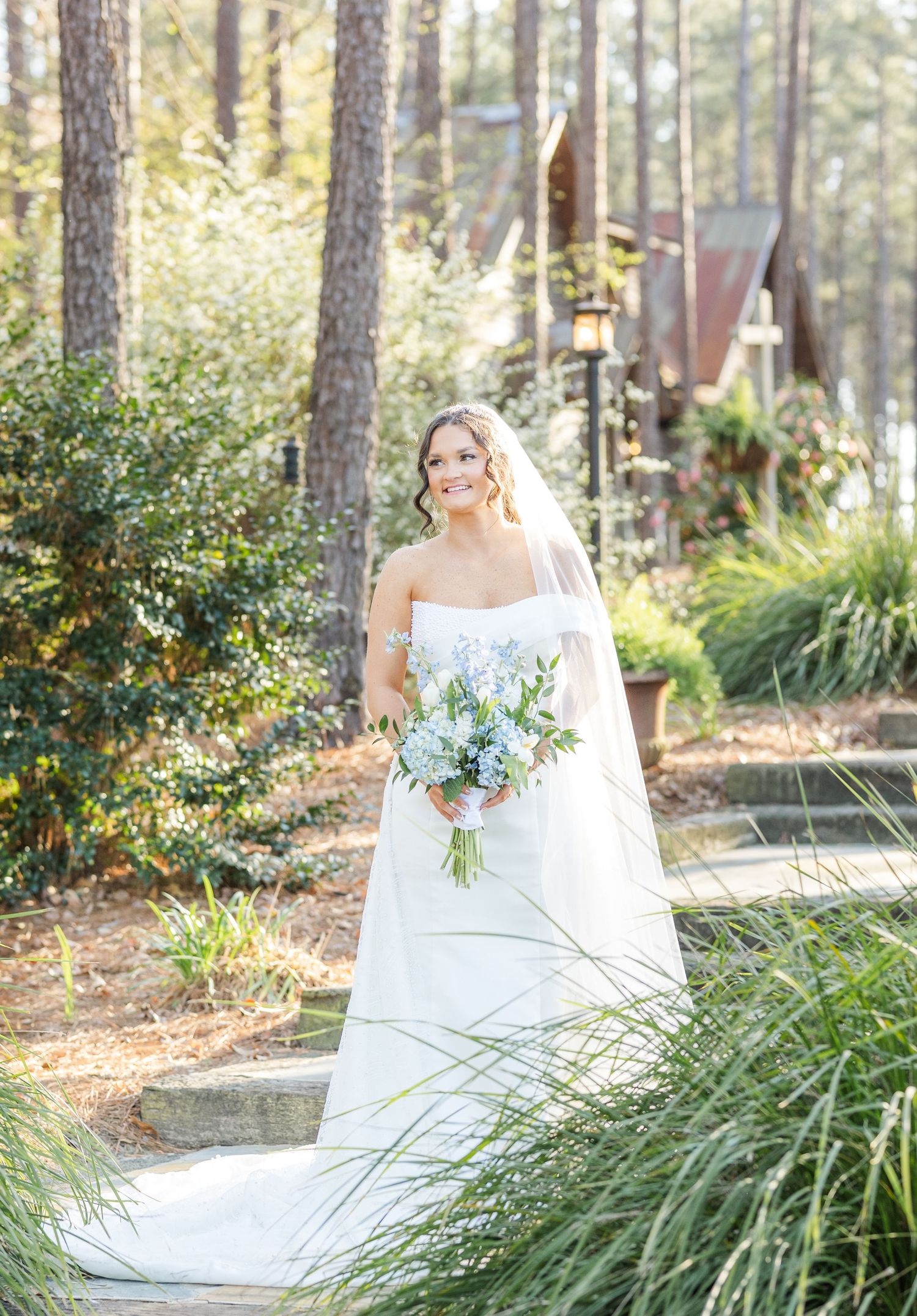 Elegant bride in strapless white gown with veil holding blue and white bouquet in sunlit pine tree forest setting.