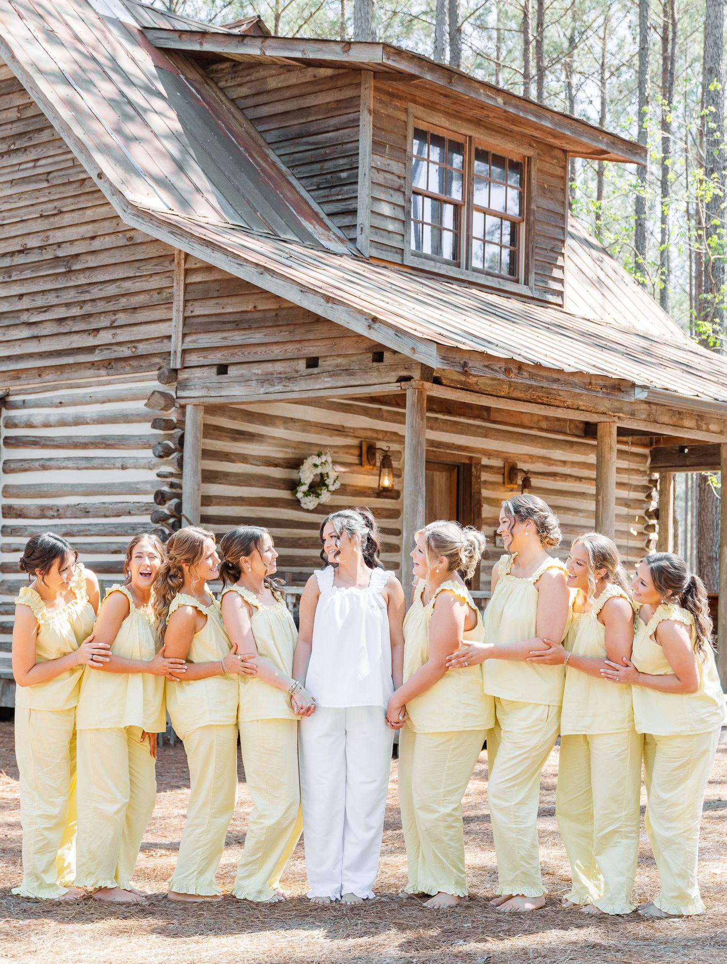 Bride in white with bridesmaids in yellow dresses standing before a rustic log cabin.