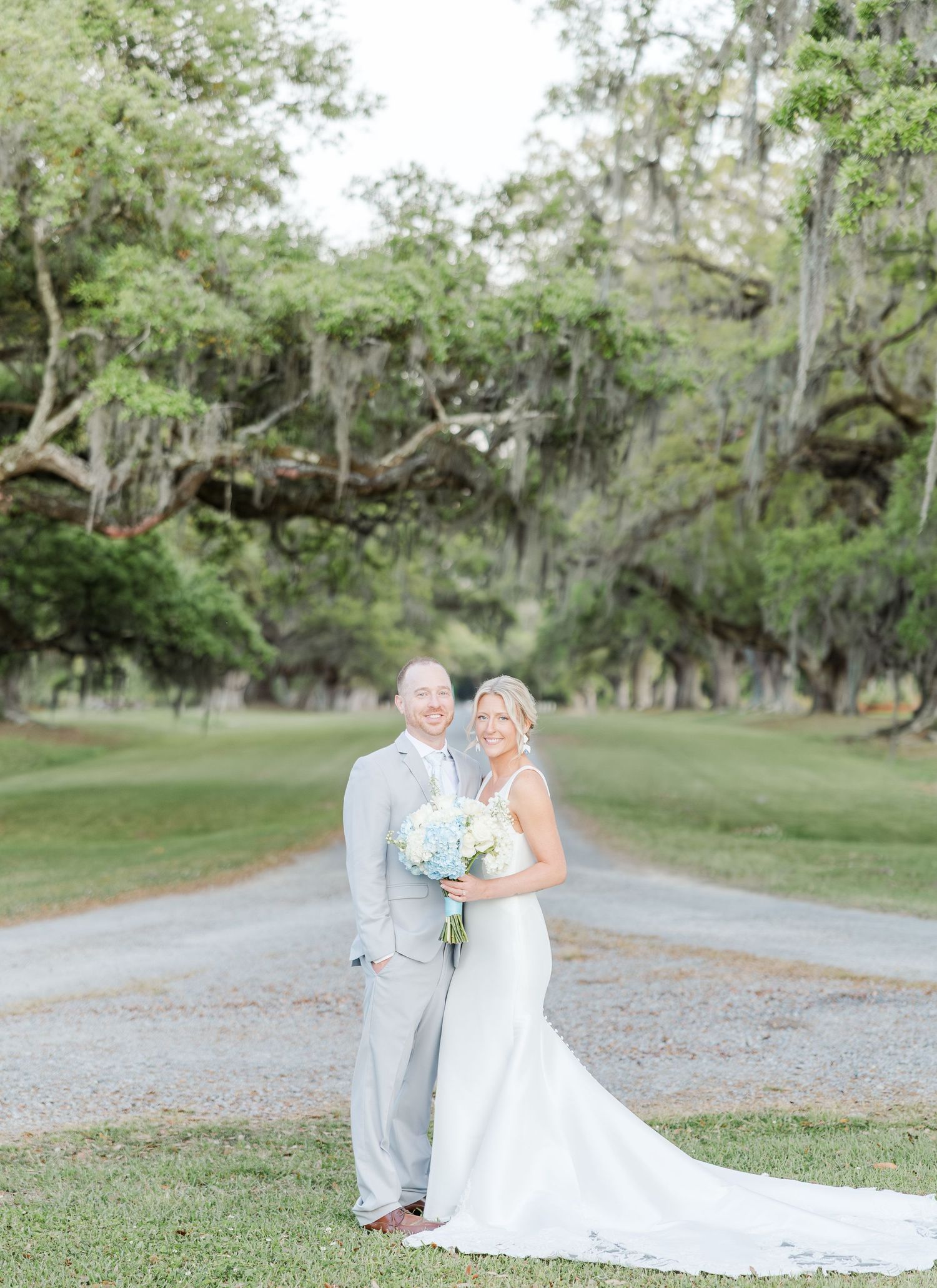 Bride and groom posing on a tree-lined gravel path, bride holding blue and white bouquet at outdoor wedding venue.
