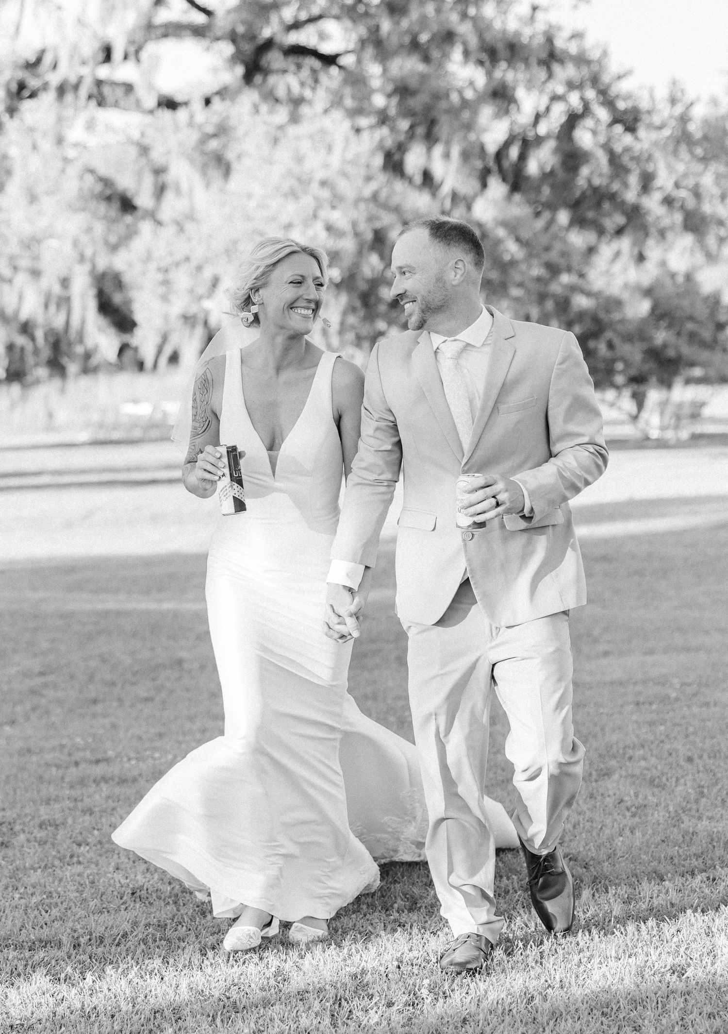 Black and white photo of smiling bride and groom walking hand in hand across a grassy lawn at their wedding.