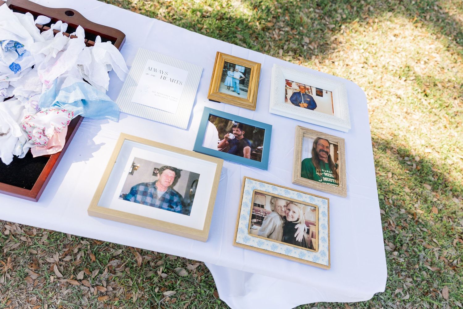 Outdoor wedding memory table displaying framed family photos, gift box with blue ribbon, and keepsakes on white tablecloth.