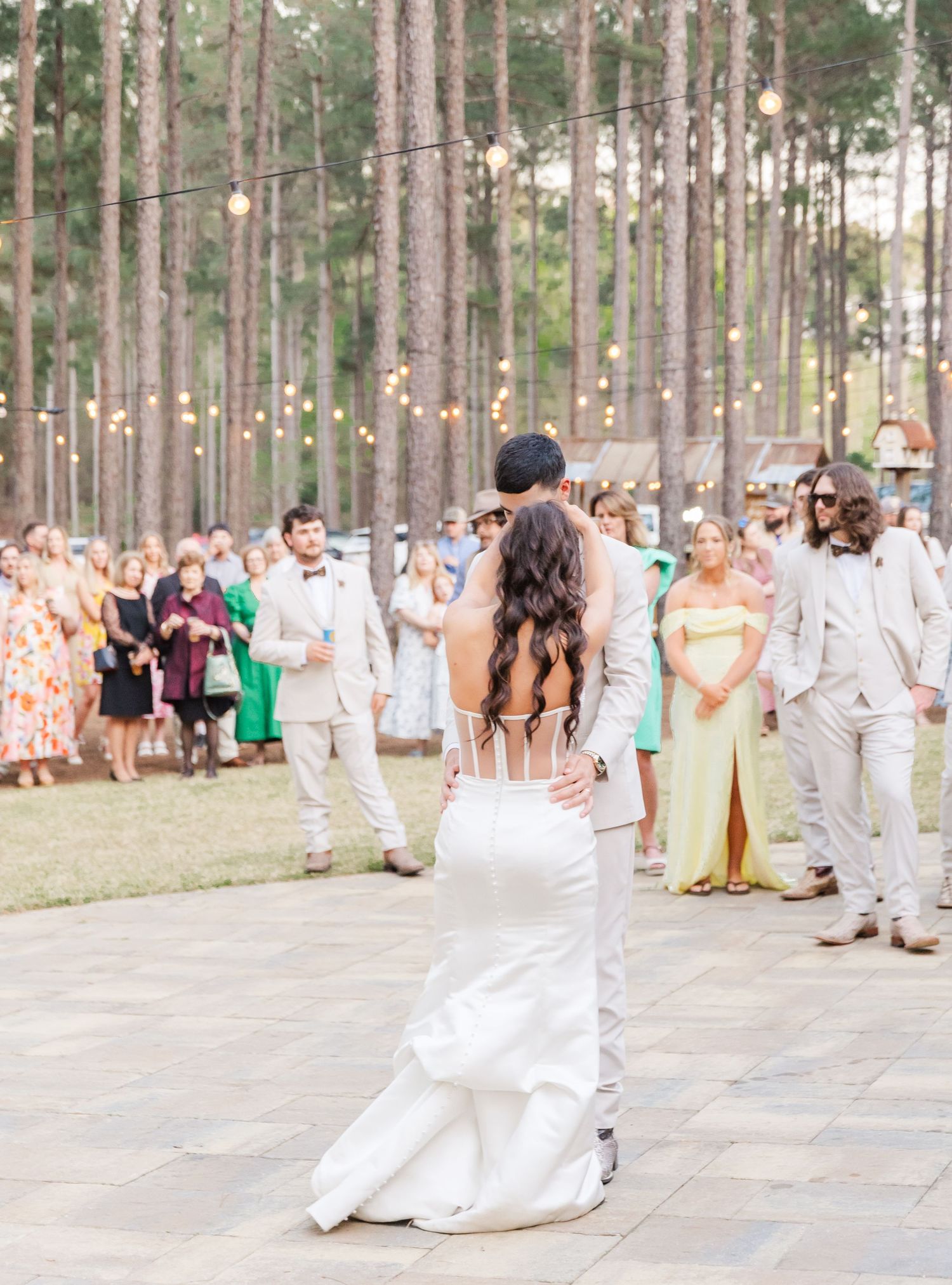 Bride and groom share first dance on outdoor patio surrounded by guests and string lights in pine forest.