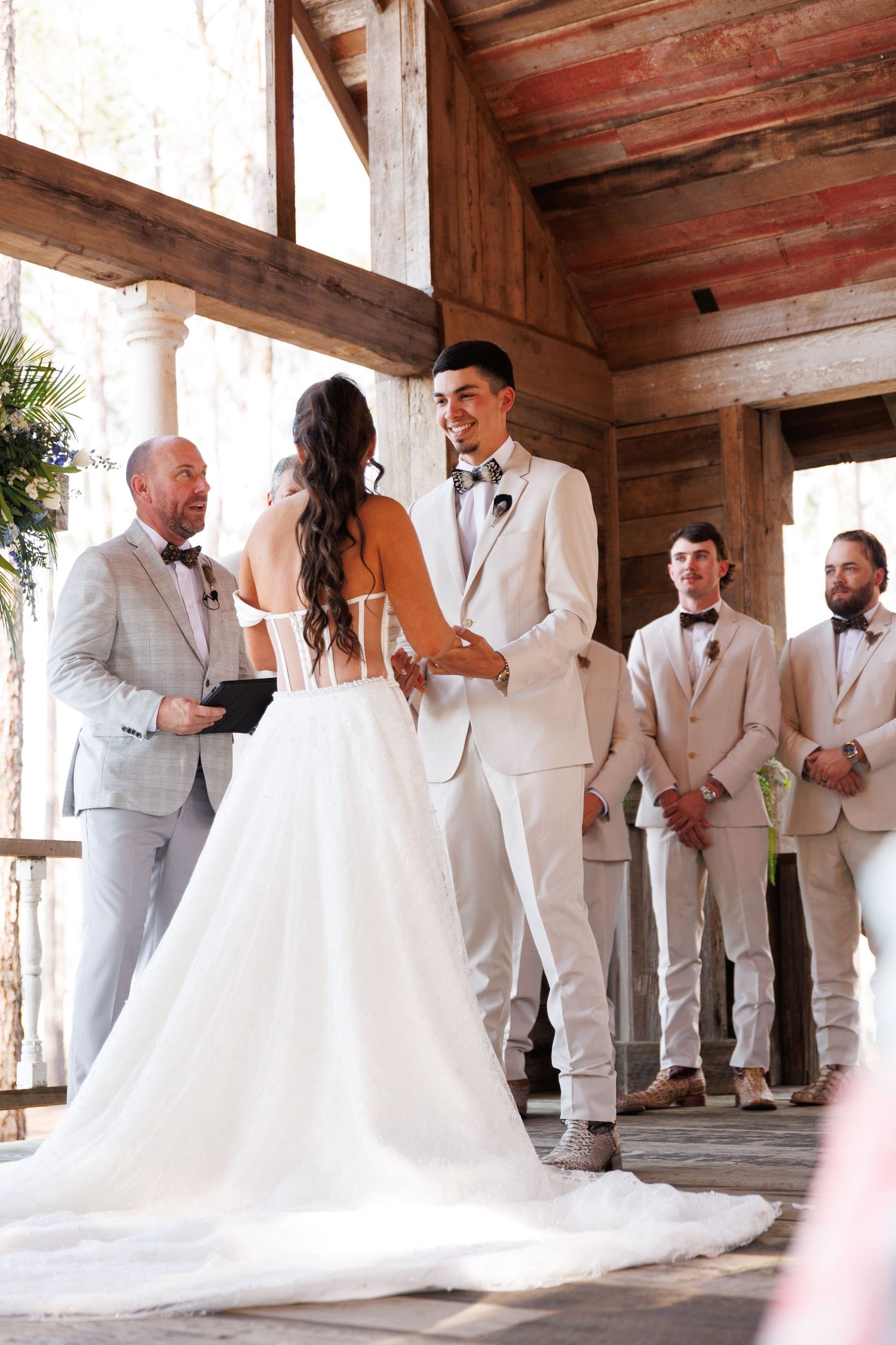 Bride and groom exchanging vows at rustic barn wedding ceremony with groomsmen standing nearby.