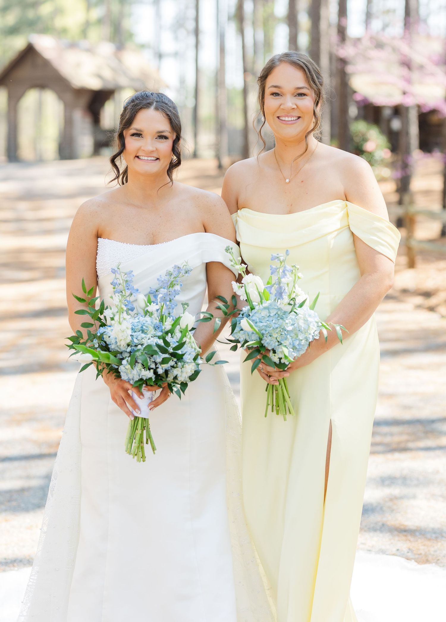 Bride in strapless white gown and bridesmaid in yellow off-shoulder dress holding blue floral bouquets.