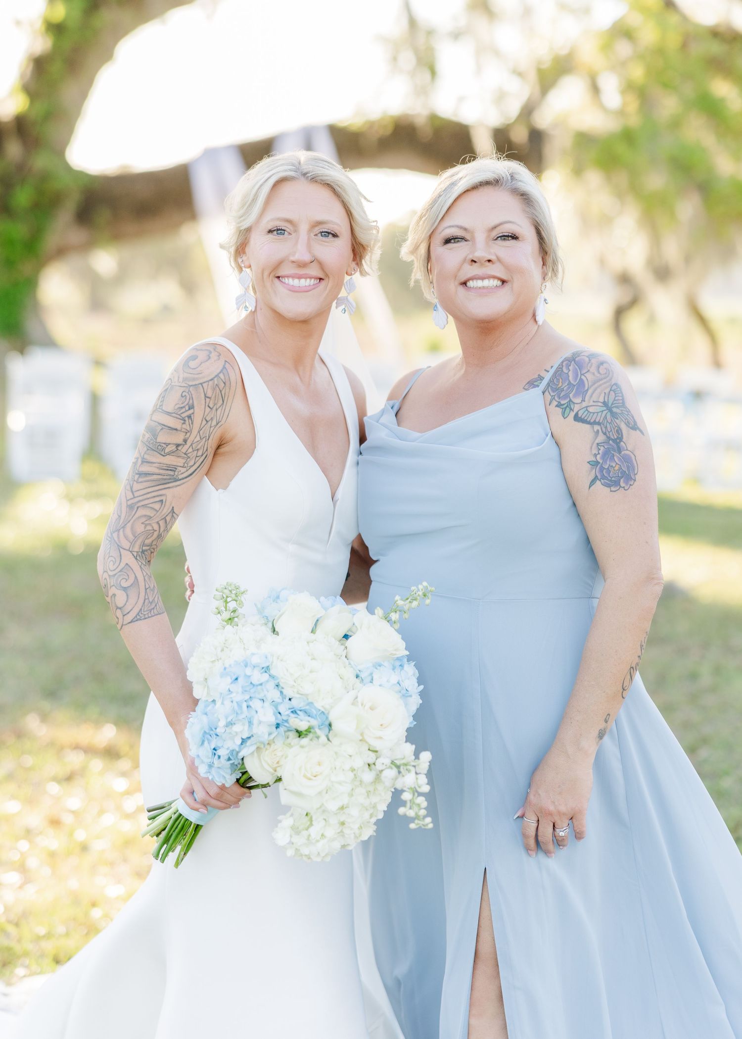 Tattooed bride in white gown poses with bridesmaid in light blue dress holding floral bouquet.