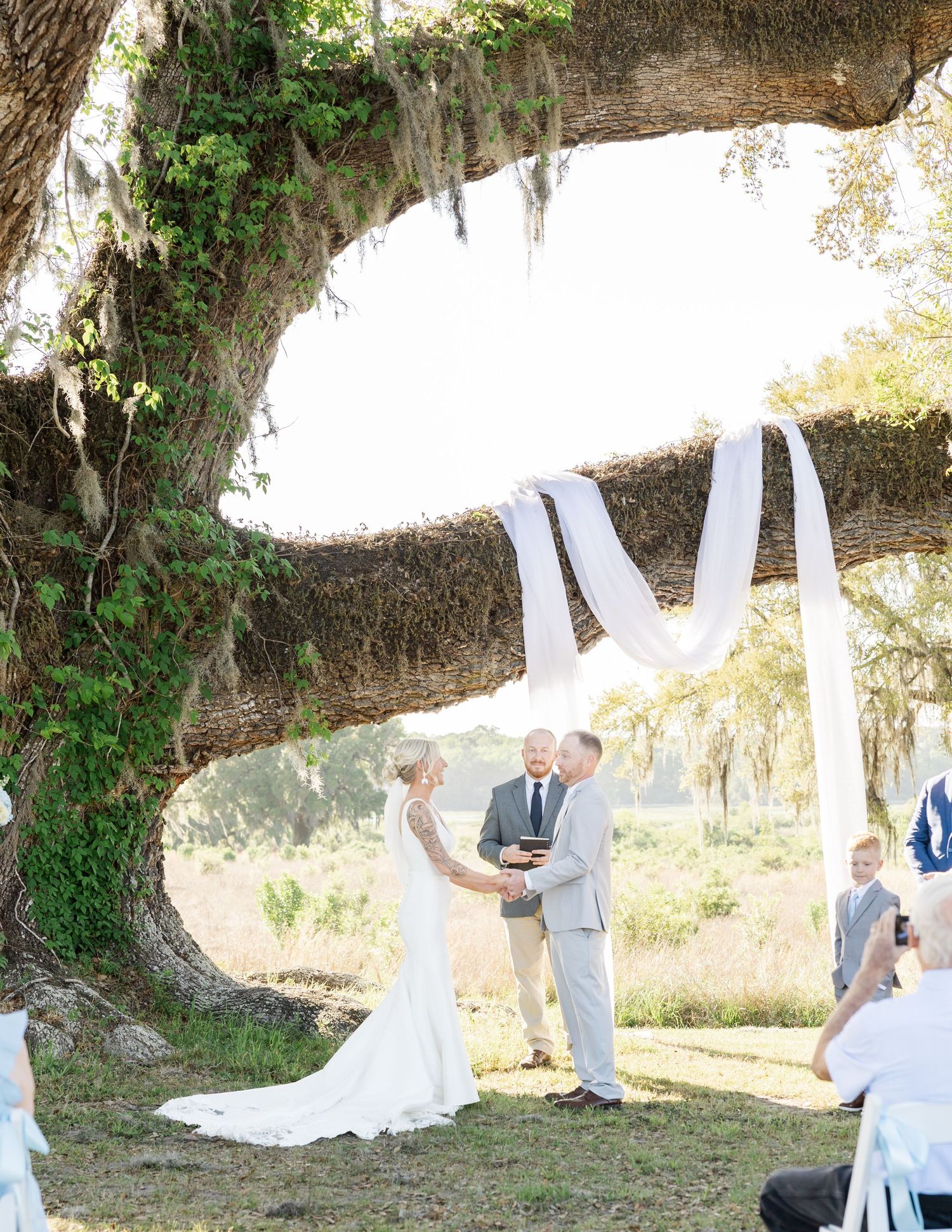 Bride and groom exchange vows under a grand oak tree with white fabric draping at outdoor ceremony.