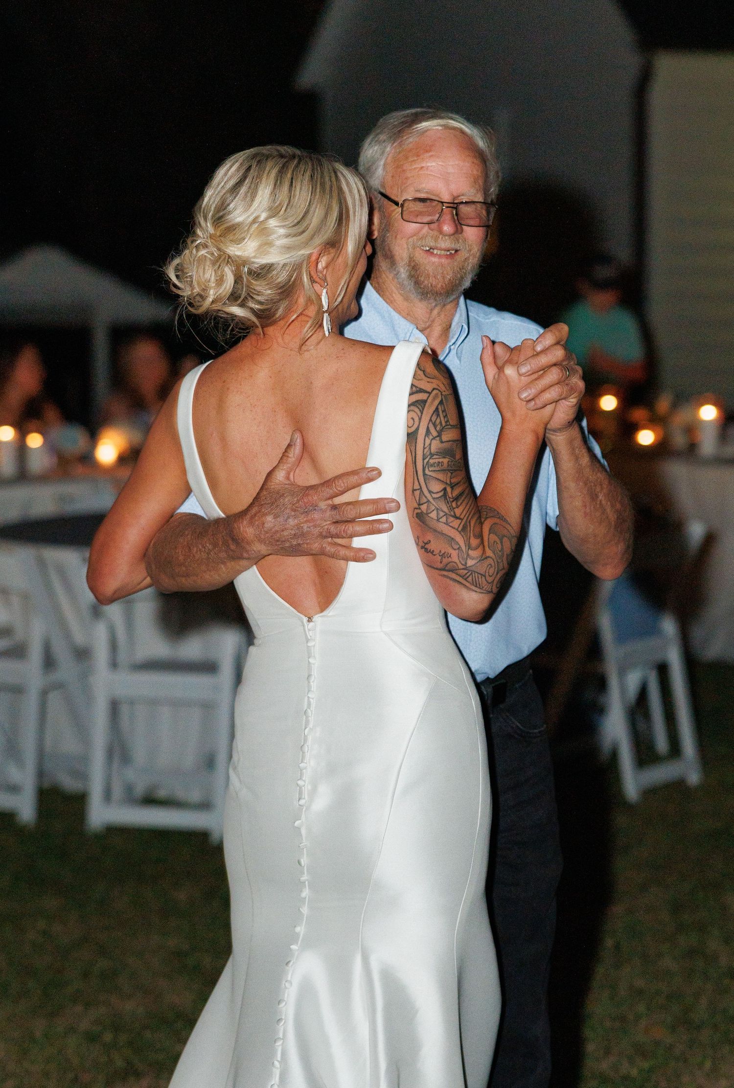 Bride in a backless white gown dancing with an older man with tattoos at an outdoor evening reception.