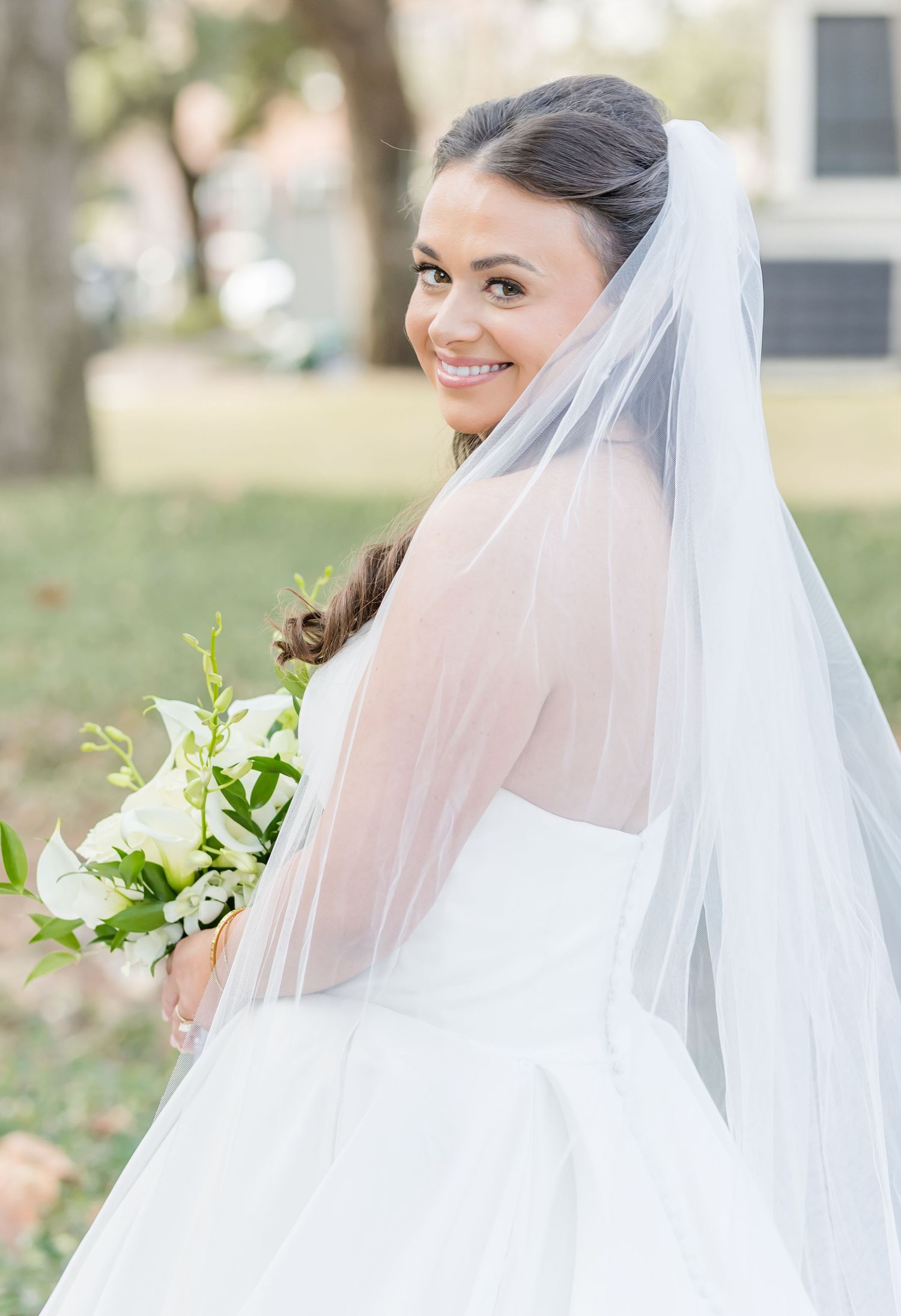 Smiling bride in white gown and veil holding white floral bouquet, posing outdoors on her wedding day.