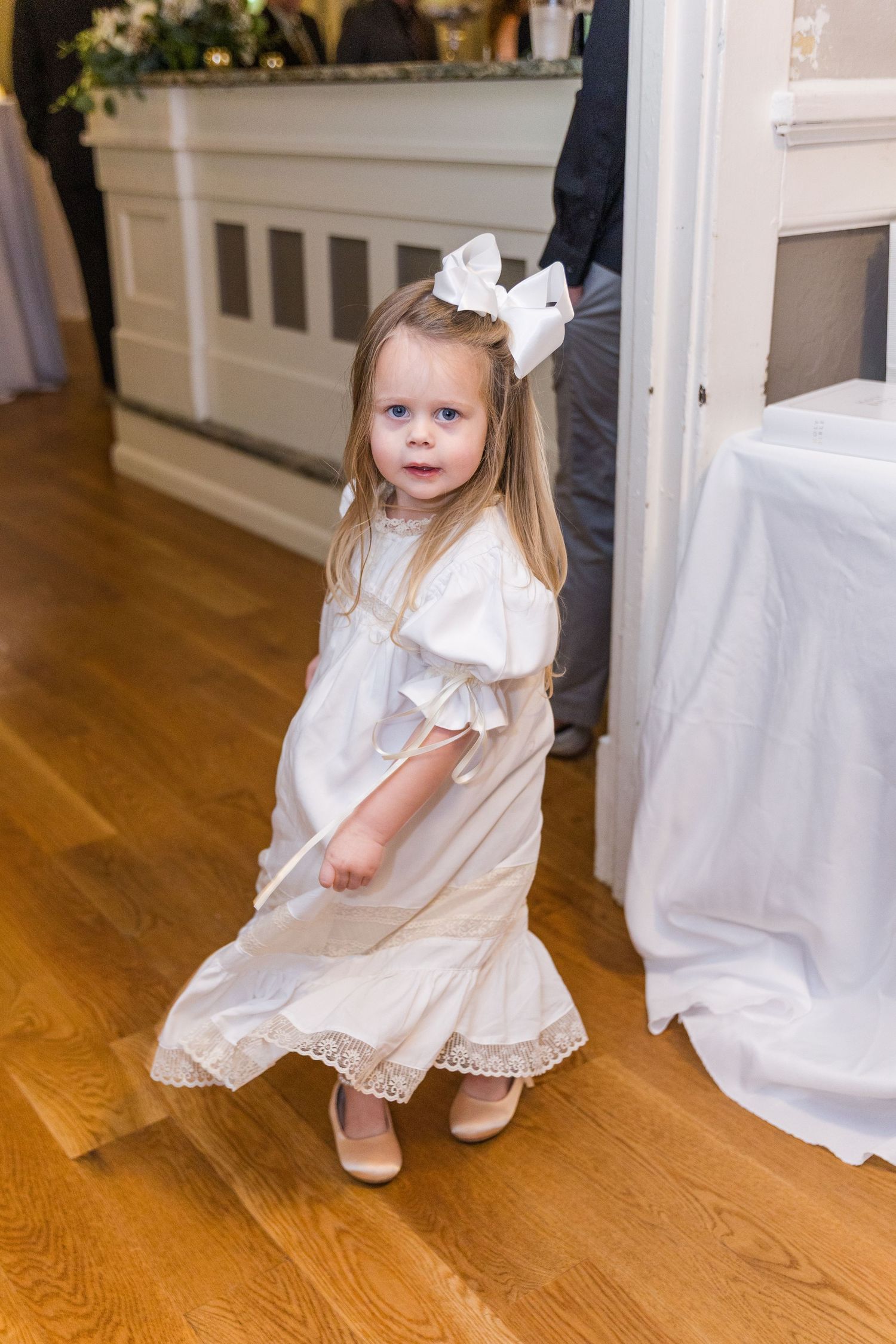 A little flower girl in a white dress and bow dances at a wedding reception on hardwood floors.